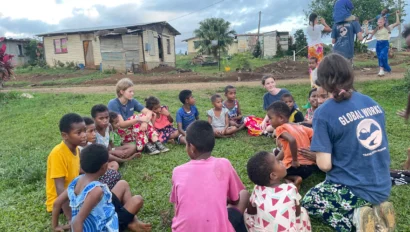 A group of children and volunteers sit on grass in a circle, surrounded by modest houses and trees, under a cloudy sky.