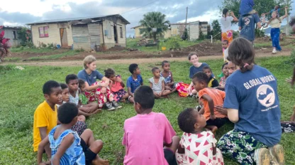 A group of children and volunteers sit on grass in a circle, surrounded by modest houses and trees, under a cloudy sky.