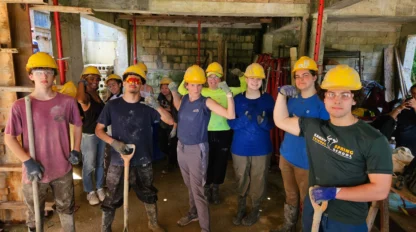 A group of people wearing yellow hard hats and work gloves, posing with tools in a construction site.