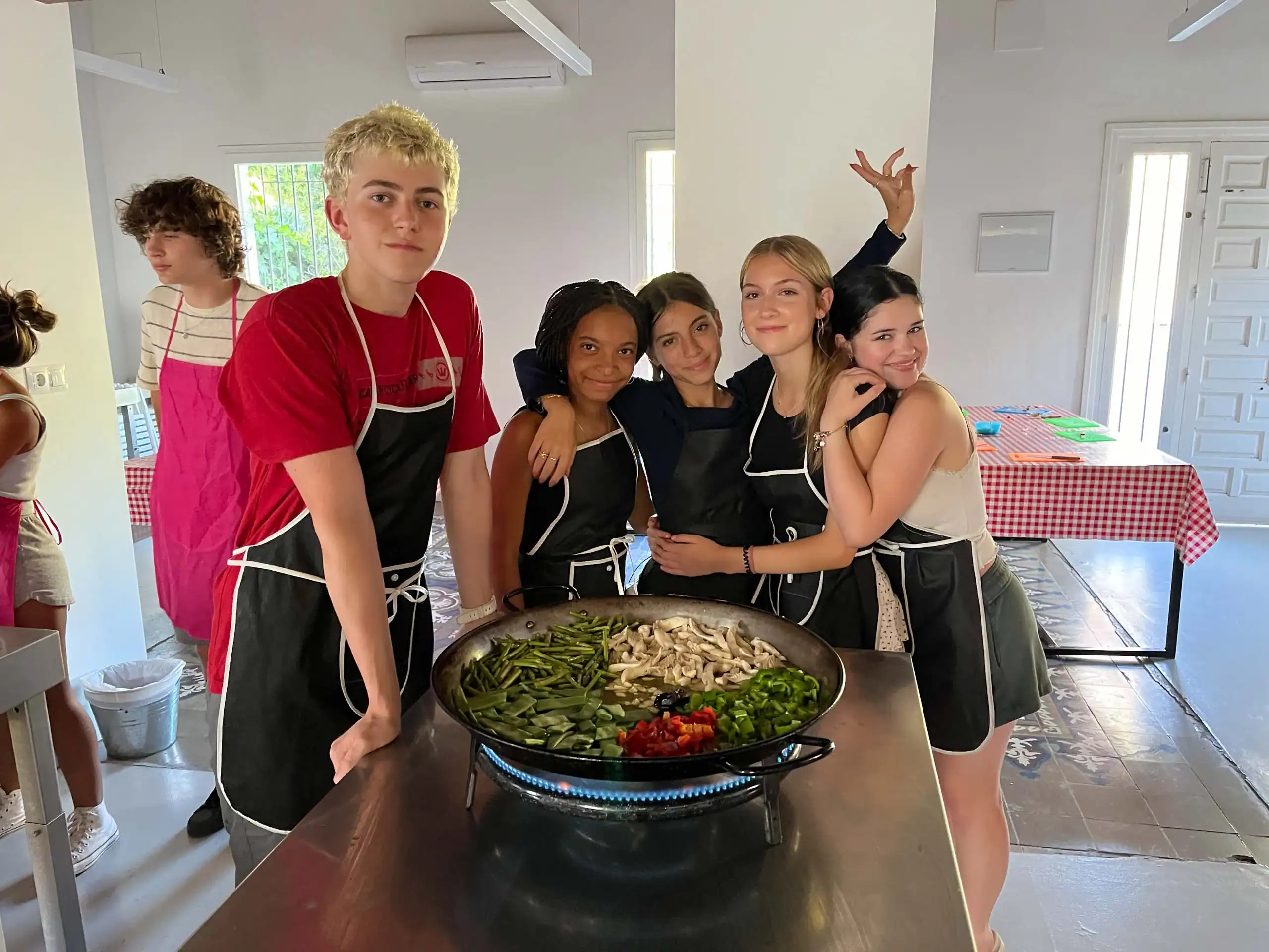 Five teenagers wearing aprons stand around a large pan of vegetables in a bright kitchen, posing for a group photo during their spanish immersion program to Spain.