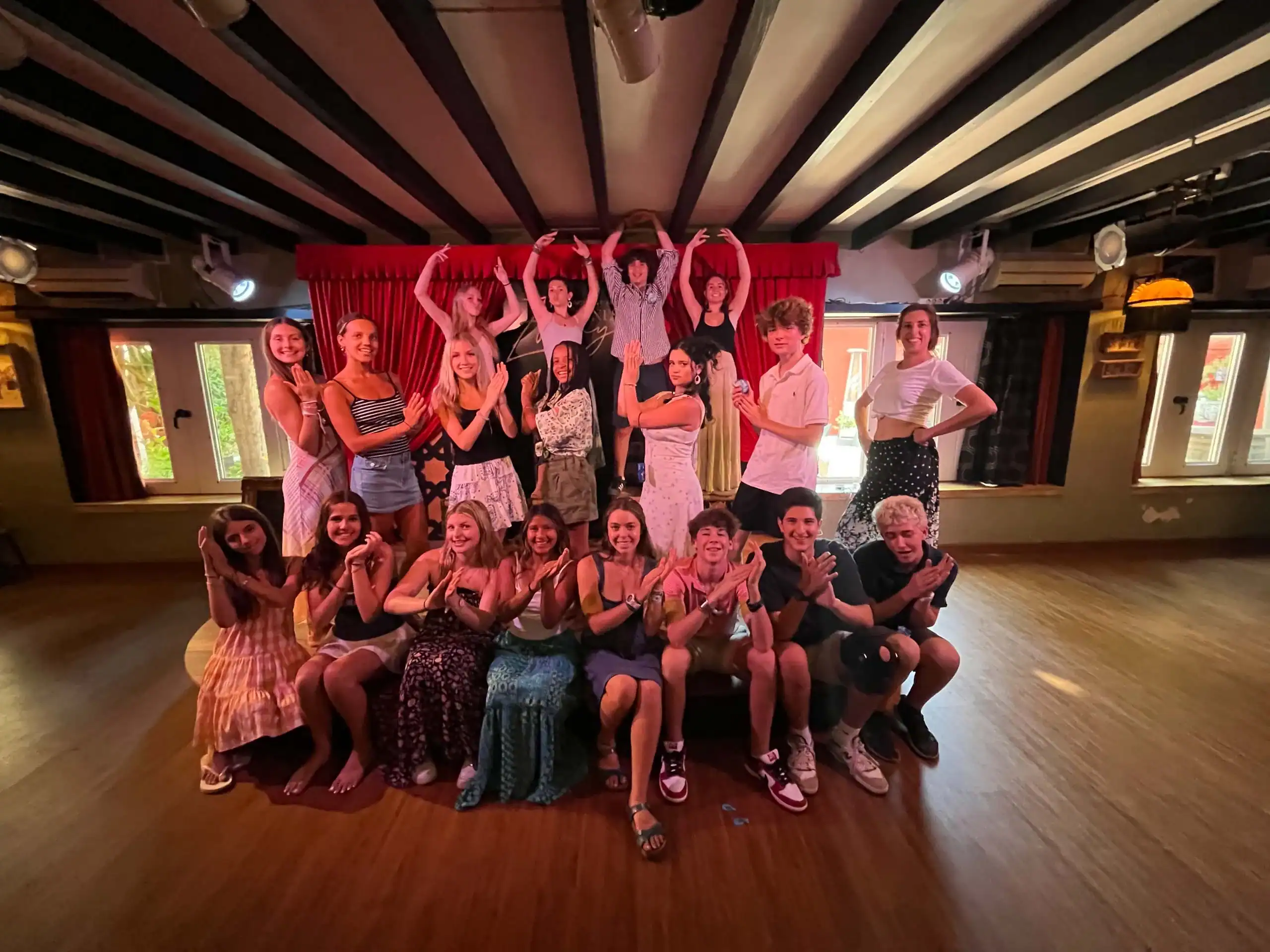 A group of sixteen people pose indoors in two rows, some sitting on the floor and others standing behind, making various hand and arm gestures during their spanish immersion program to Spain.