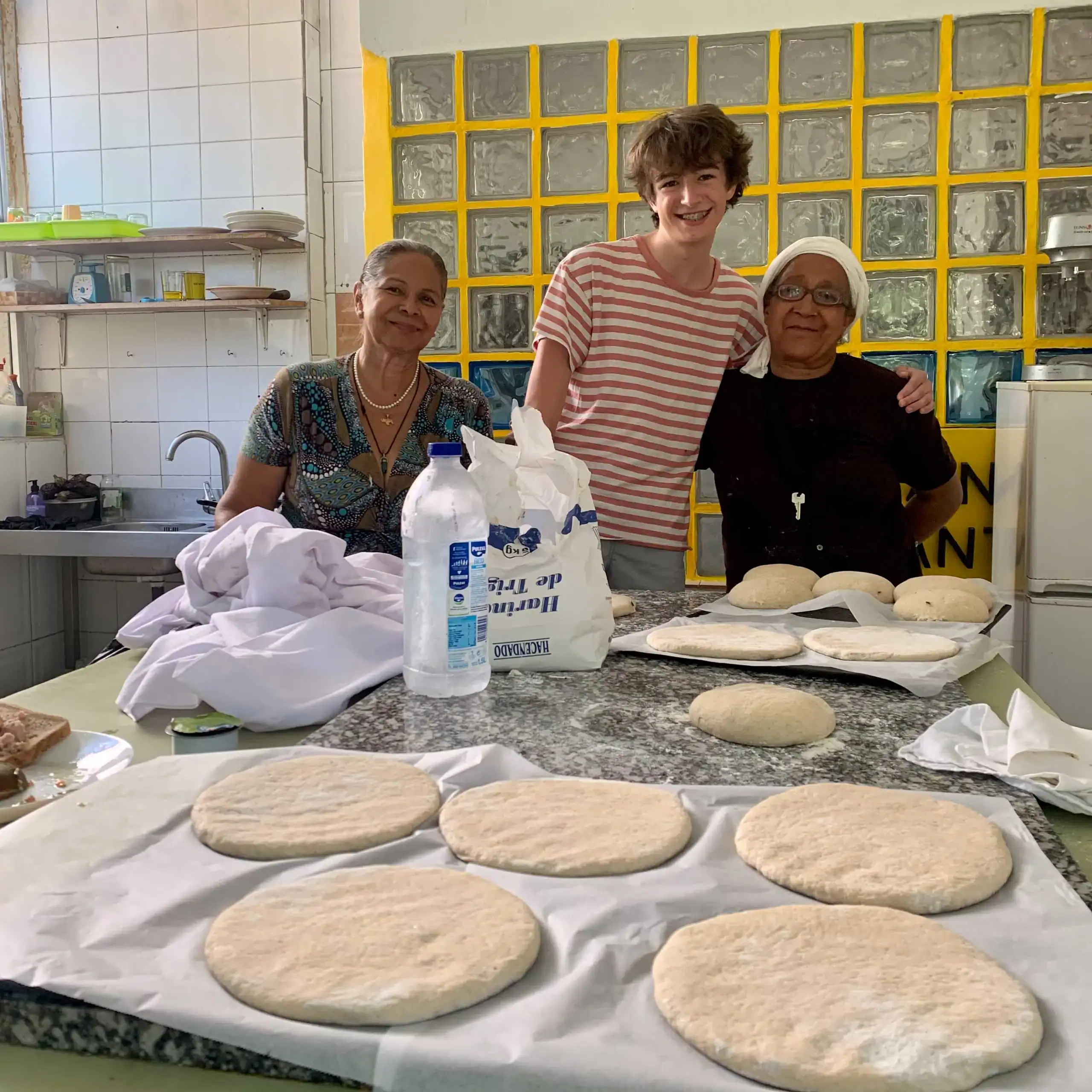 Three people stand in a kitchen behind a counter with dough rounds on baking sheets, preparing bread during their Spanish immersion program.