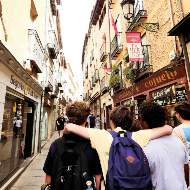 A group of teenagers with backpacks walk down a narrow street lined with shops and balconies in a European town, enjoying their spanish immersion program to spain.