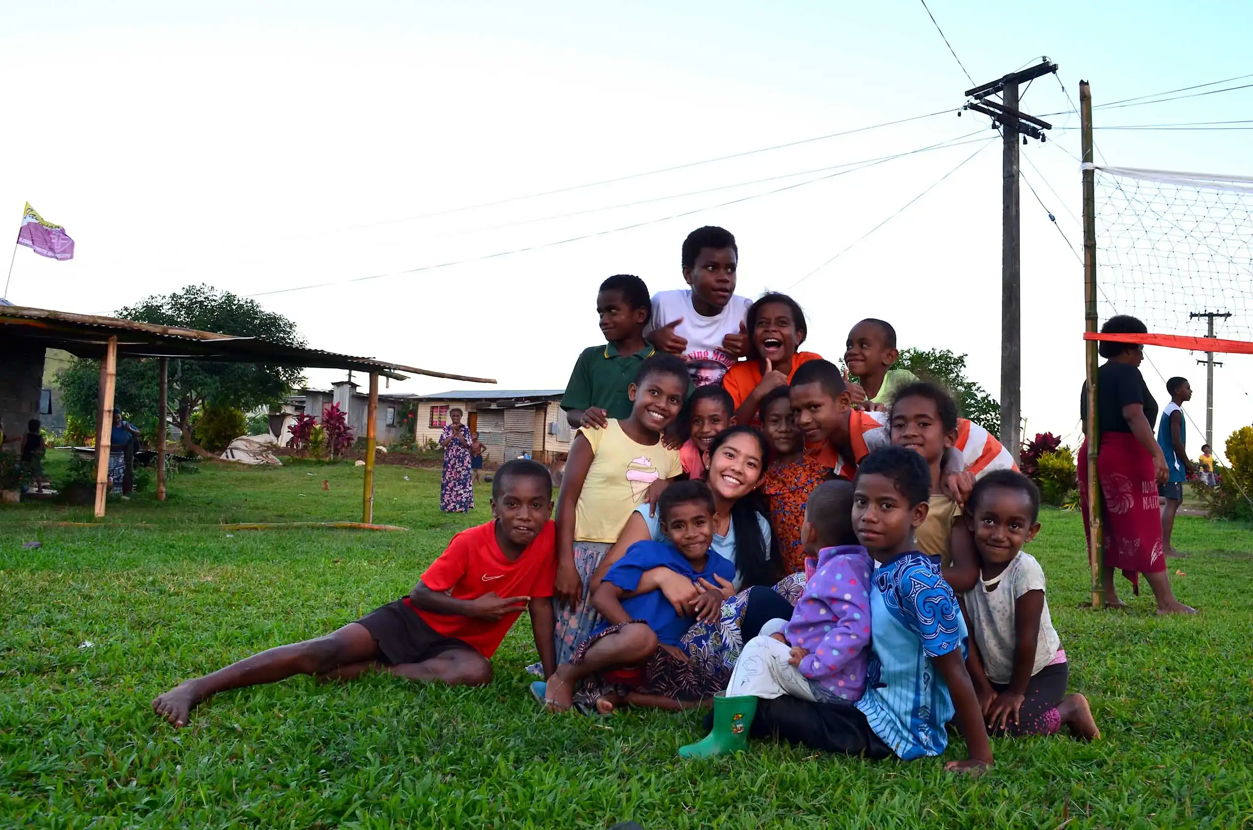 A group of children and an adult, part of a summer service program, sit and kneel together on green grass, smiling at the camera in an outdoor setting with buildings and utility poles in the background.