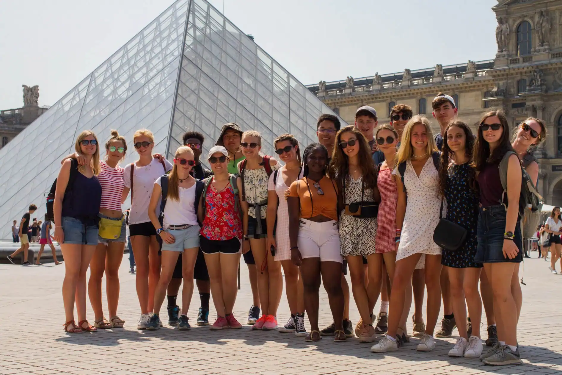 A group of young people on a student trip to France poses for a photo in front of the Louvre Pyramid in Paris on a sunny day.