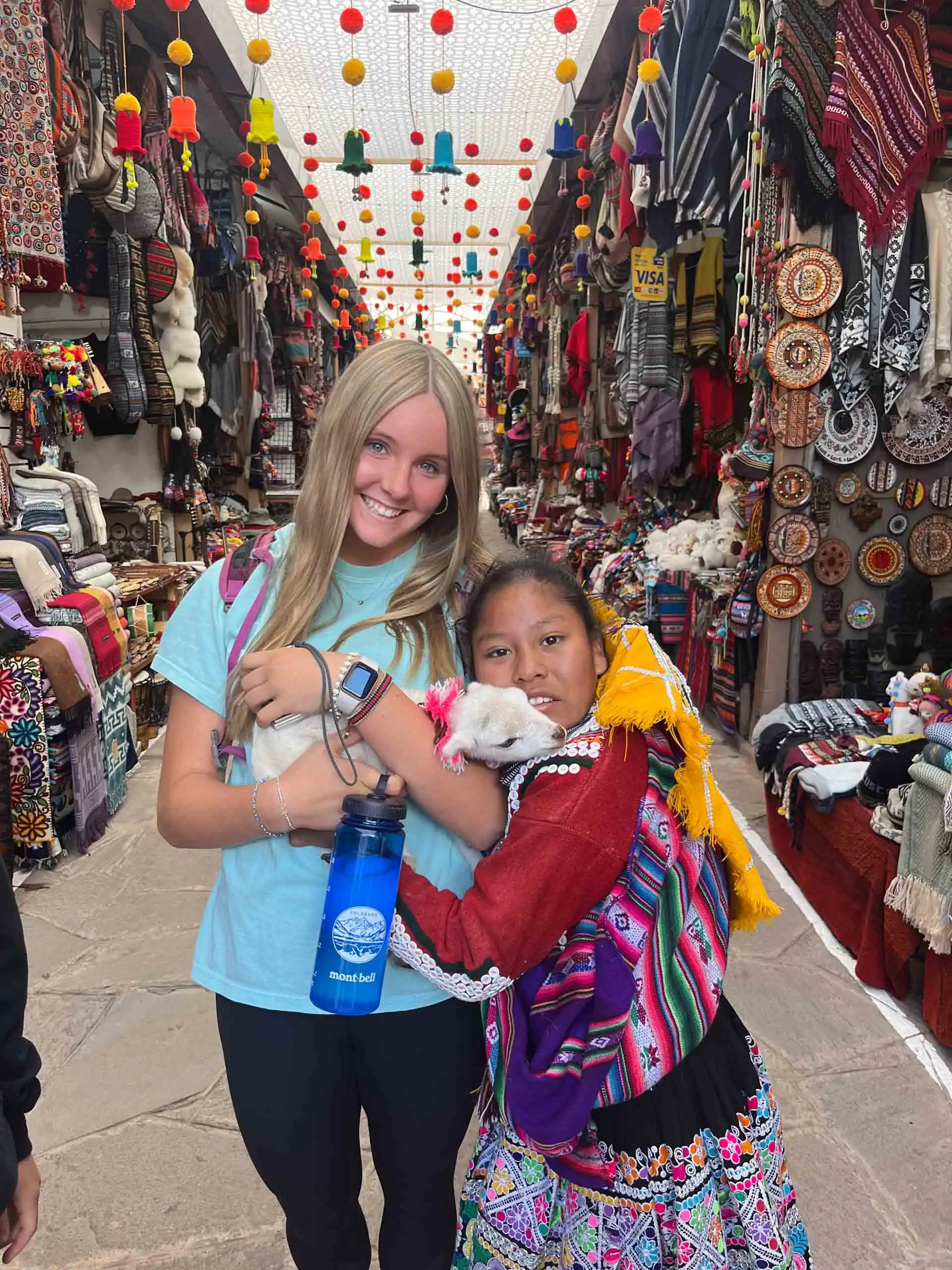 Two people smile on a student trip to Peru while holding a small white llama in a colorful market filled with textiles and crafts.