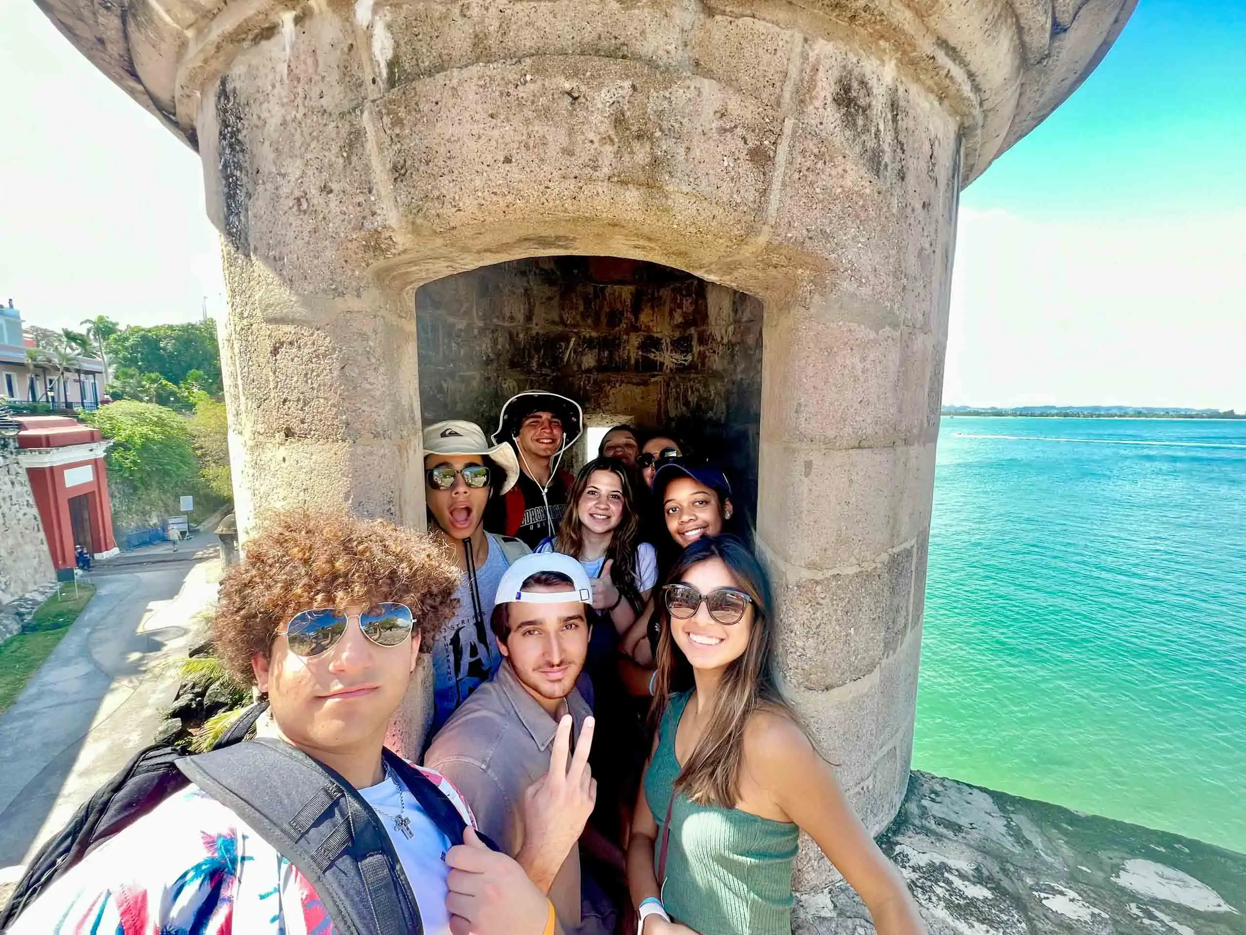 A group of young adults on a student travel to Puerto Rico pose for a photo inside a stone turret at El Morro by the waterfront on a sunny day.