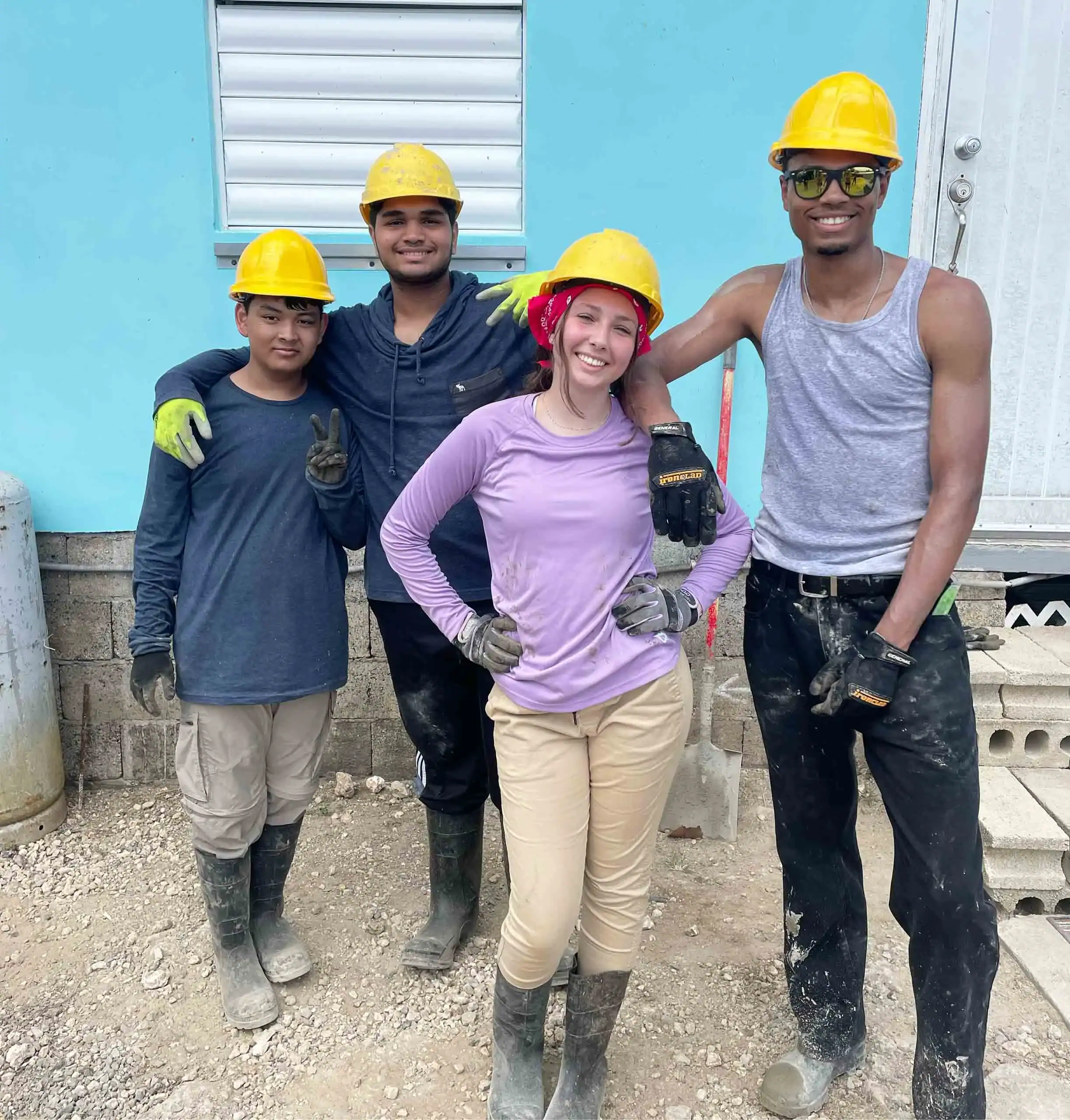 Four students wearing hard hats, gloves, and boots stand together outside a building, posing and smiling for the camera during a teen service trip to Puerto Rico.