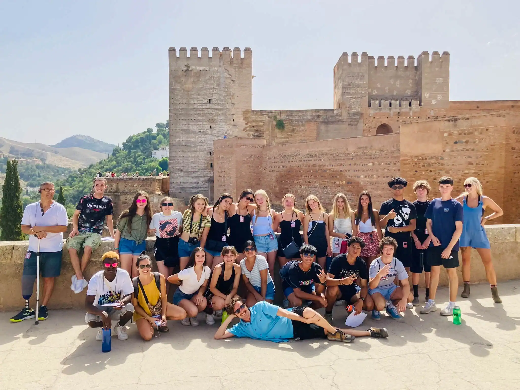 A large group of young people in a Spanish immersion program to Spain pose together in front of ancient stone walls and towers on a sunny day, with mountains visible in the background.