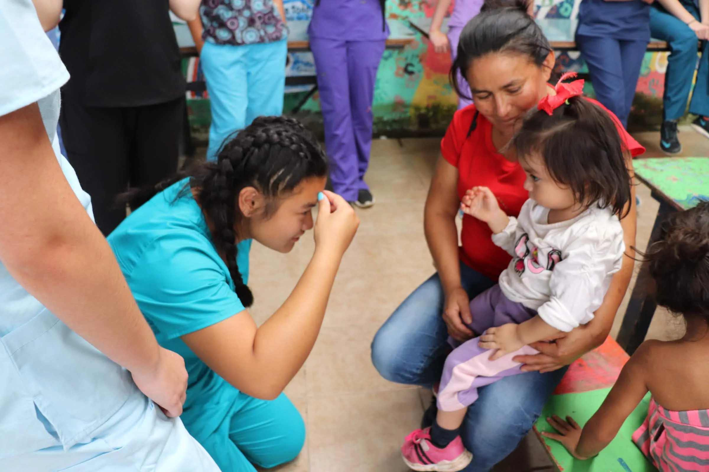 A healthcare worker in scrubs examines a young girl sitting on a woman's lap while others observe—a lively scene from a teen medical trip abroad.