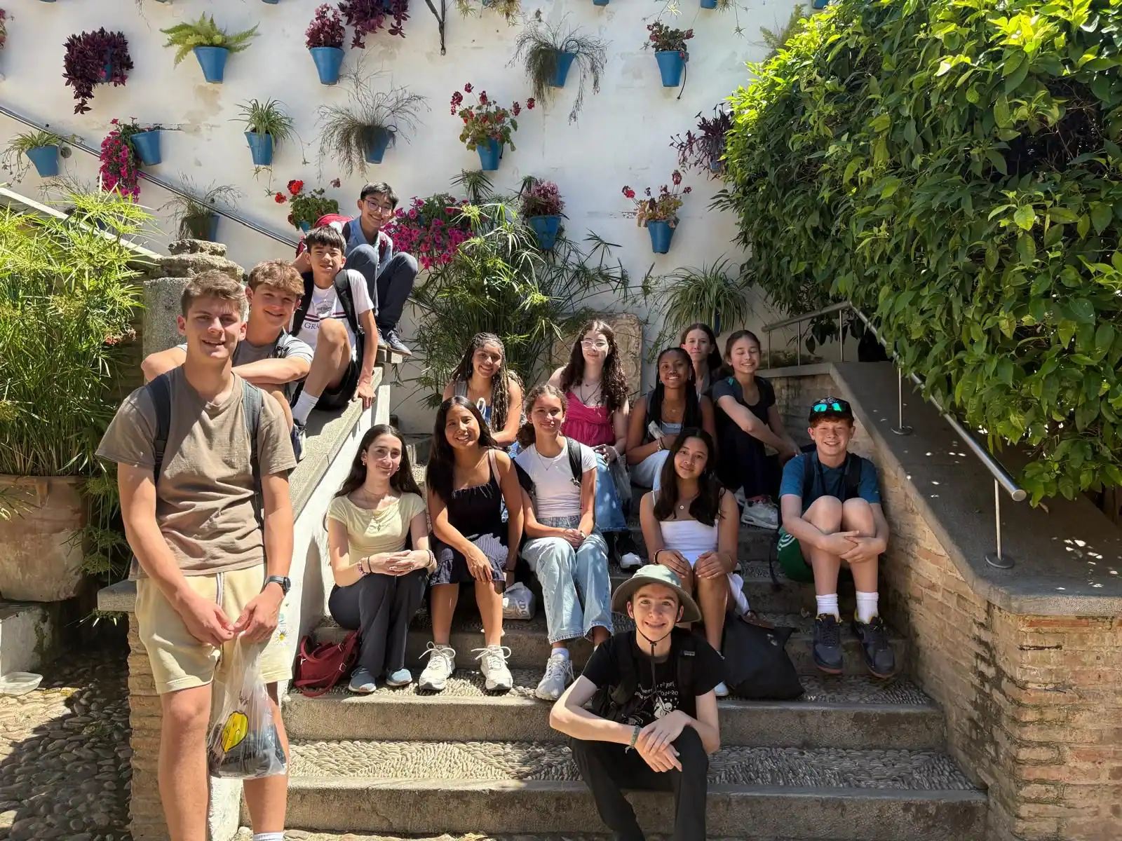A group of teenagers sits and stands on outdoor stone steps surrounded by greenery and potted plants attached to a white wall.