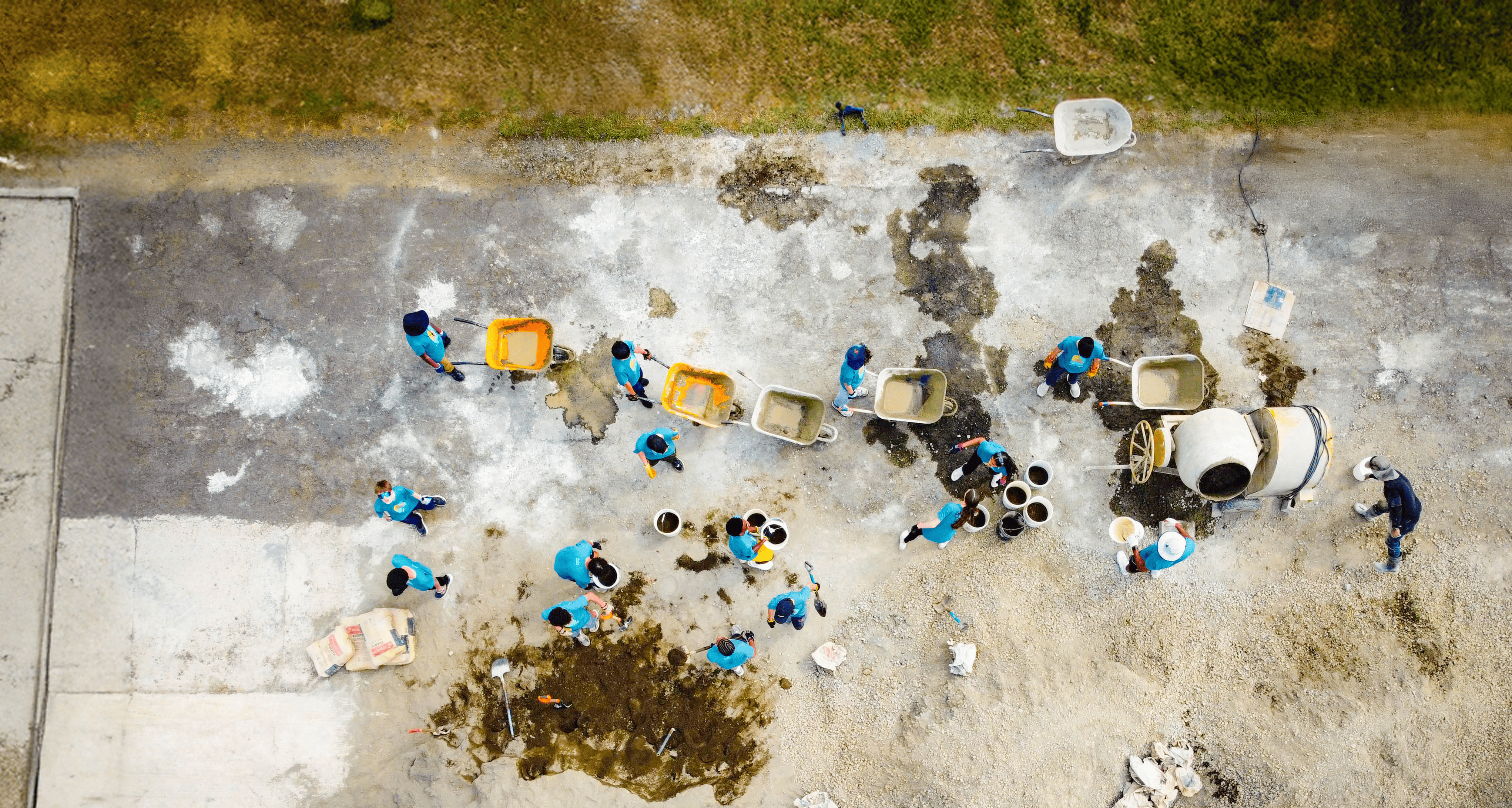 Aerial view of students in blue shirts on a school trip to Puerto Rico, working with construction materials, mixing cement, and carrying buckets on a concrete surface during a service trip.
