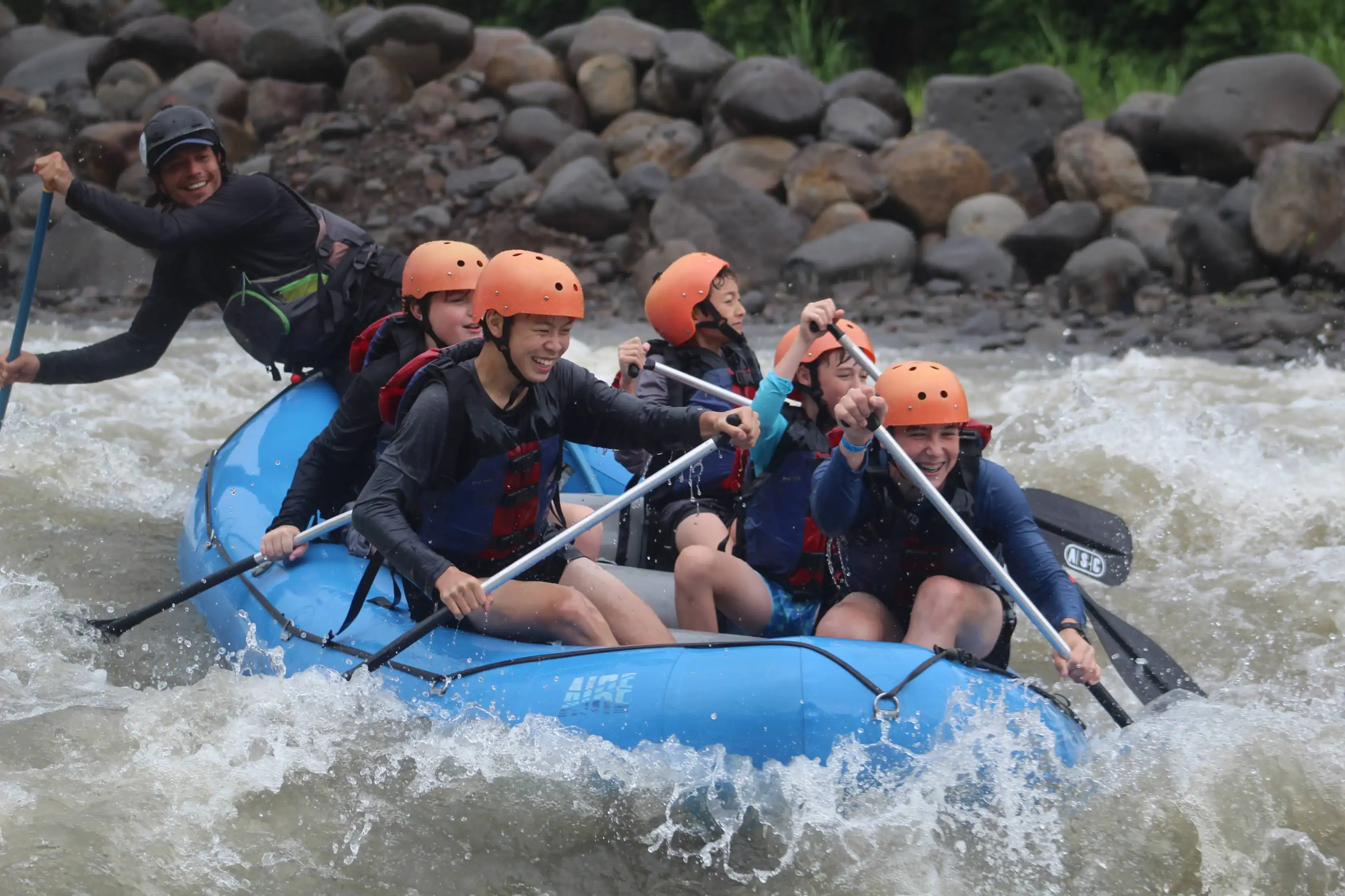 Six students wearing helmets and life jackets paddle a blue inflatable raft through whitewater rapids near a rocky riverbank, an experience often featured on Middle School student trips.
