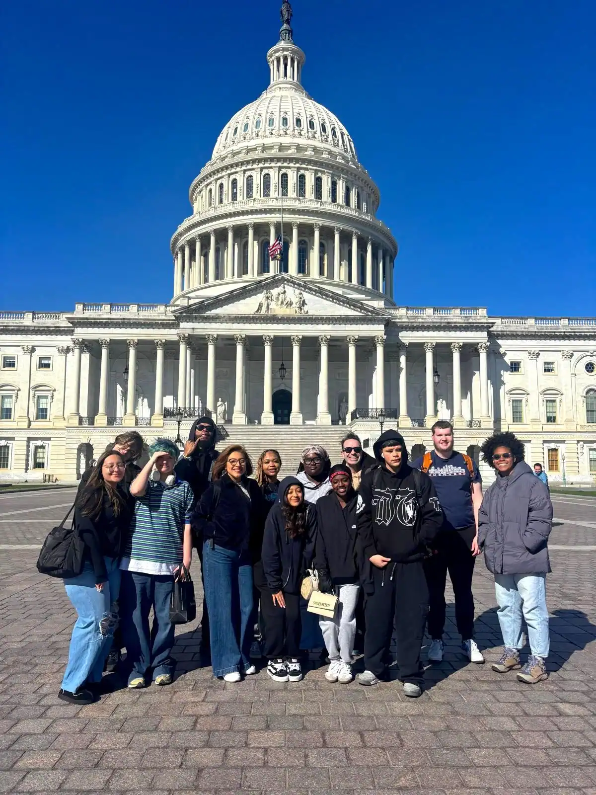 A school group travel outing captures a group of students posing for a photo in front of the United States Capitol building on a sunny day.