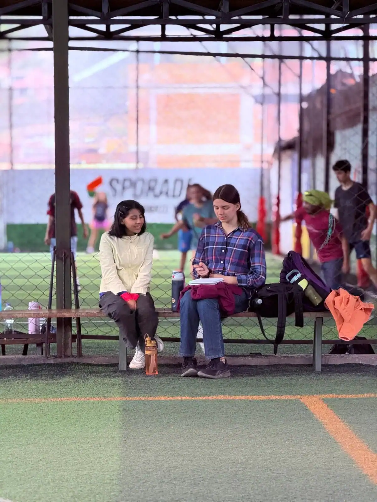 Two high school student sit on a bench under a shelter by a fenced sports field, chatting about their Spanish immersion trip. Sports bags and water bottles are beside them while people play soccer in the background.