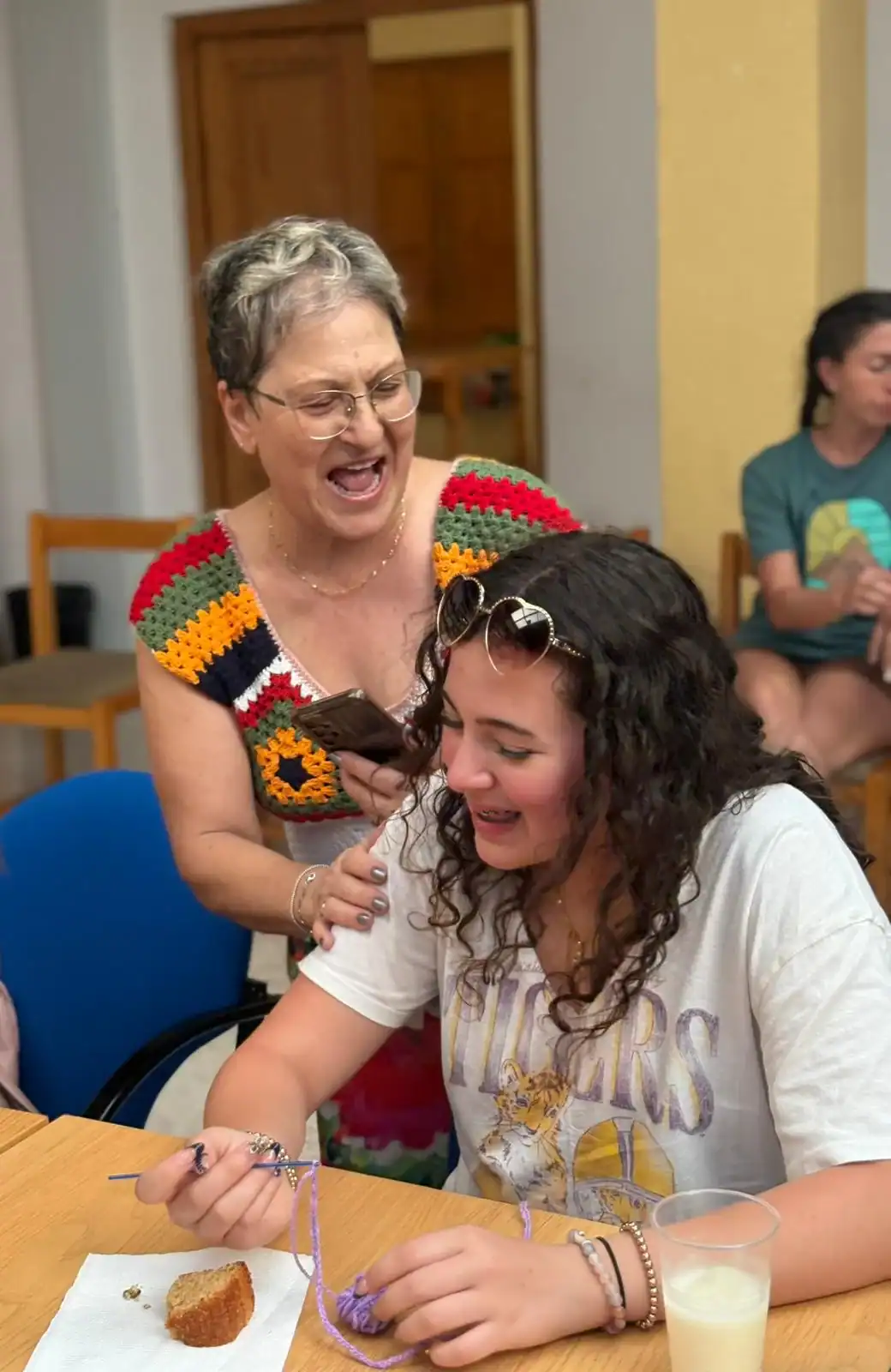 An older woman laughs and places her hand on the shoulder of a smiling high school student seated at a table, enjoying a moment together during a Global Works Spanish immersion trip, with others visible in the background.