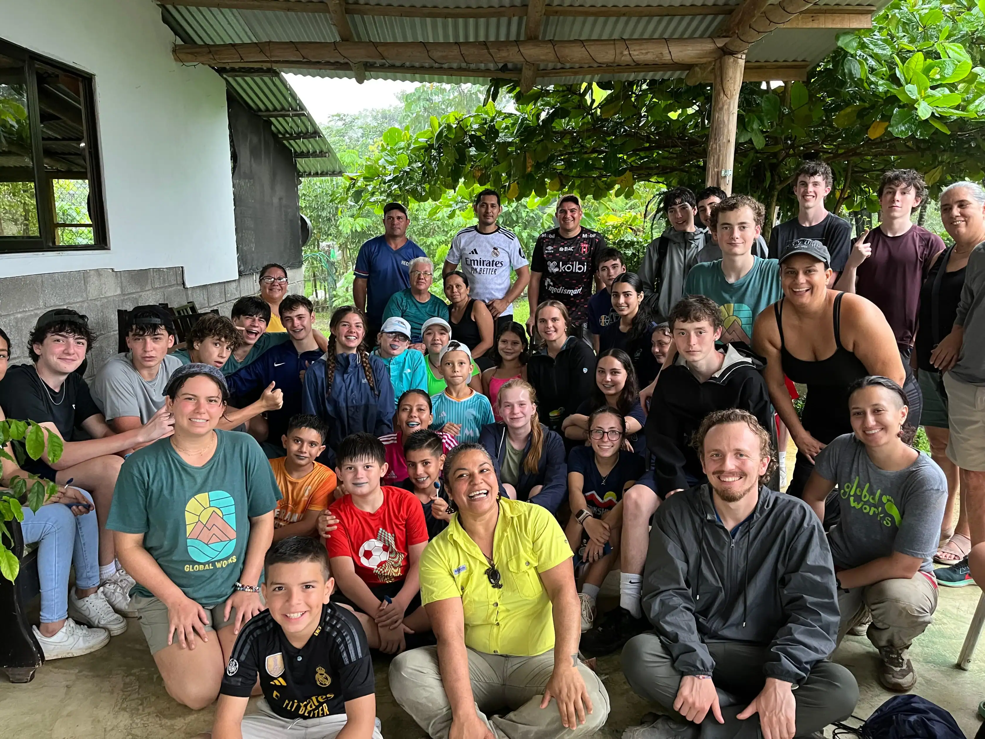 A large group of people, including adults and children, pose together on a covered patio in a tropical outdoor setting, enjoying one of the tech-free travel programs designed to foster real connections.