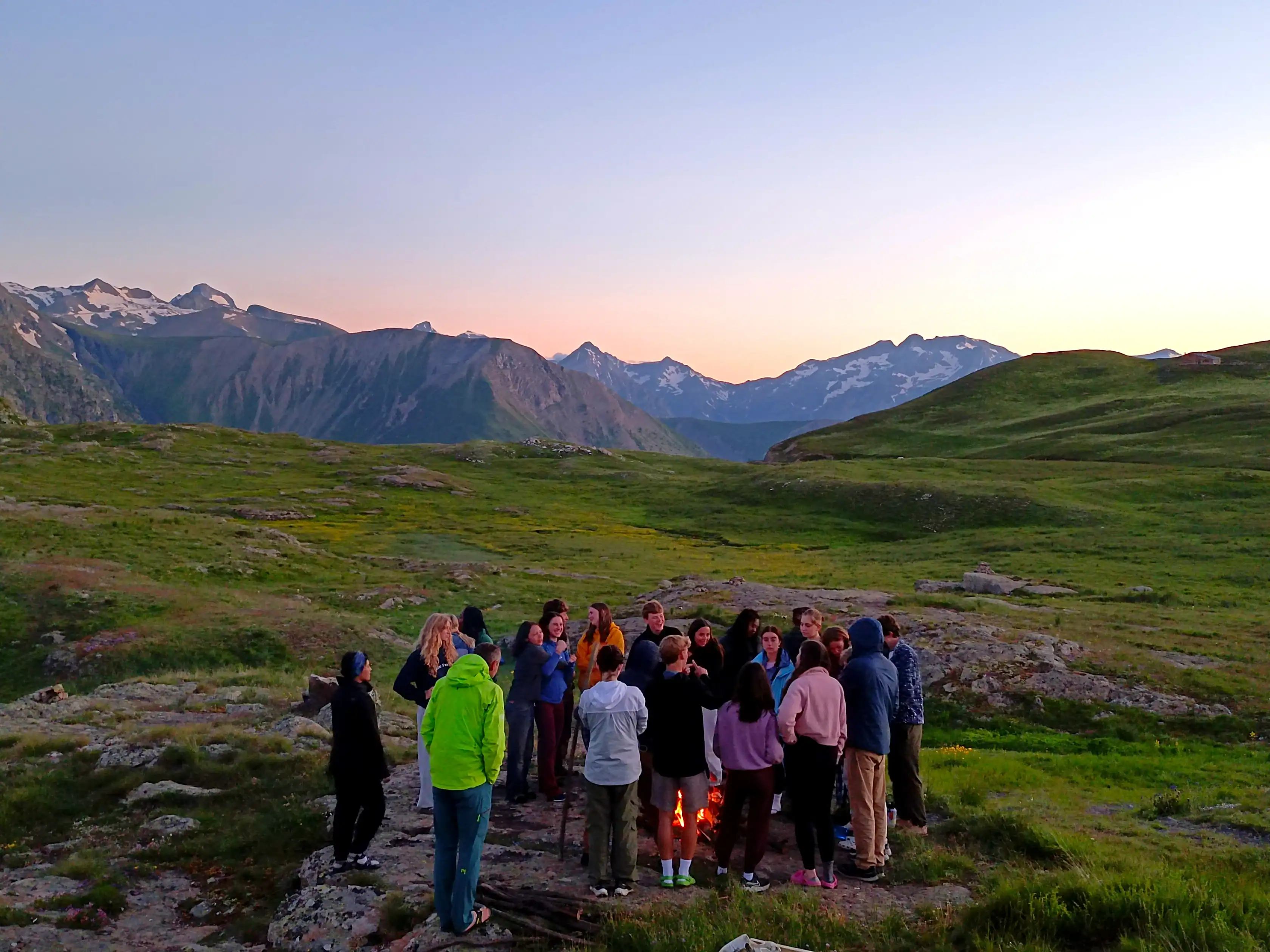 A group of people stands around a campfire on a grassy hillside with mountains and snow in the background at sunset, enjoying one of the Tech-Free travel programs that encourage unplugging and connecting with nature.