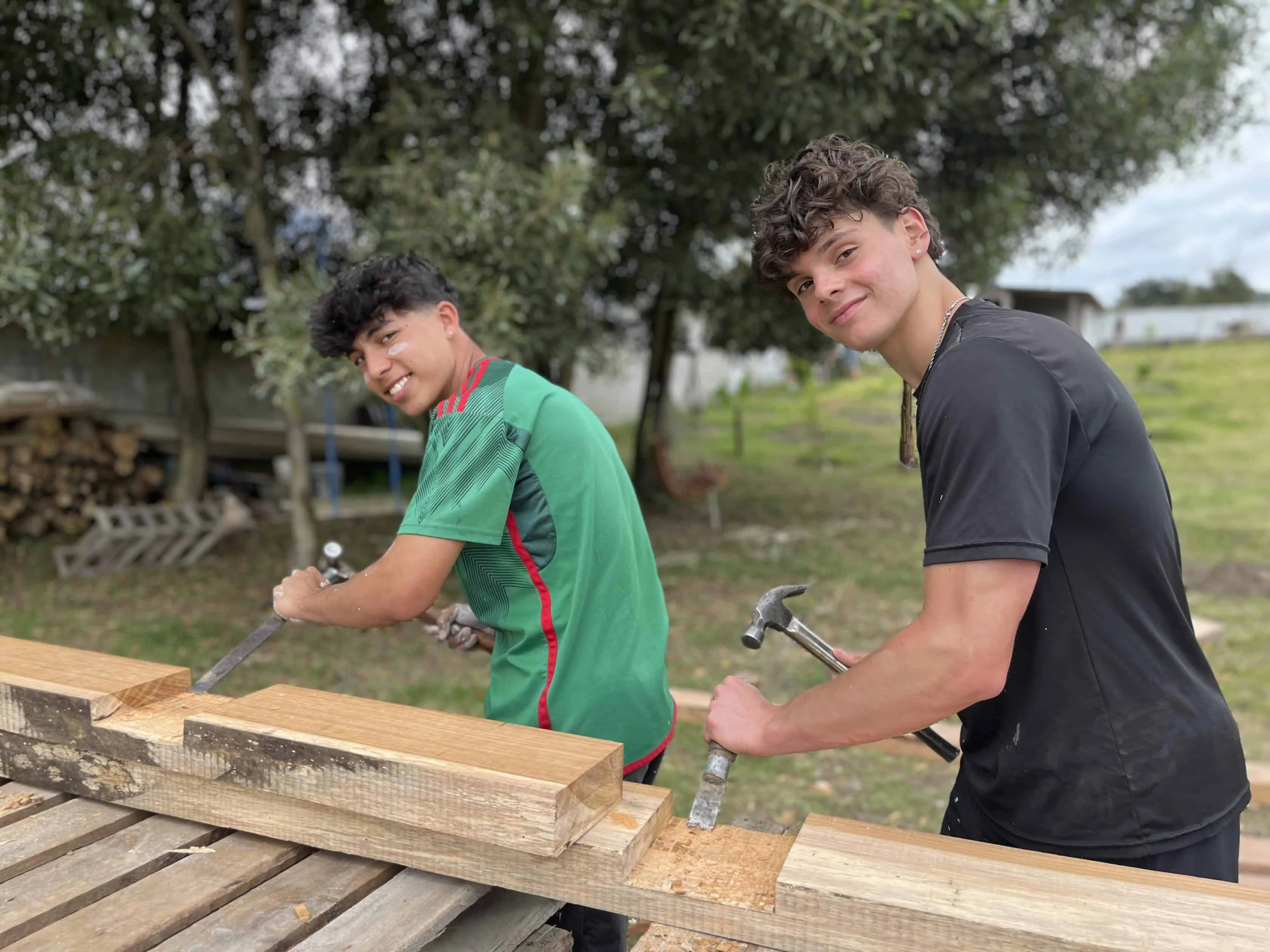 Two young men work outdoors, using chisels and hammers on wooden beams, with trees and stacked wood in the background—a scene reminiscent of hands-on experiences found in teen travel programs.