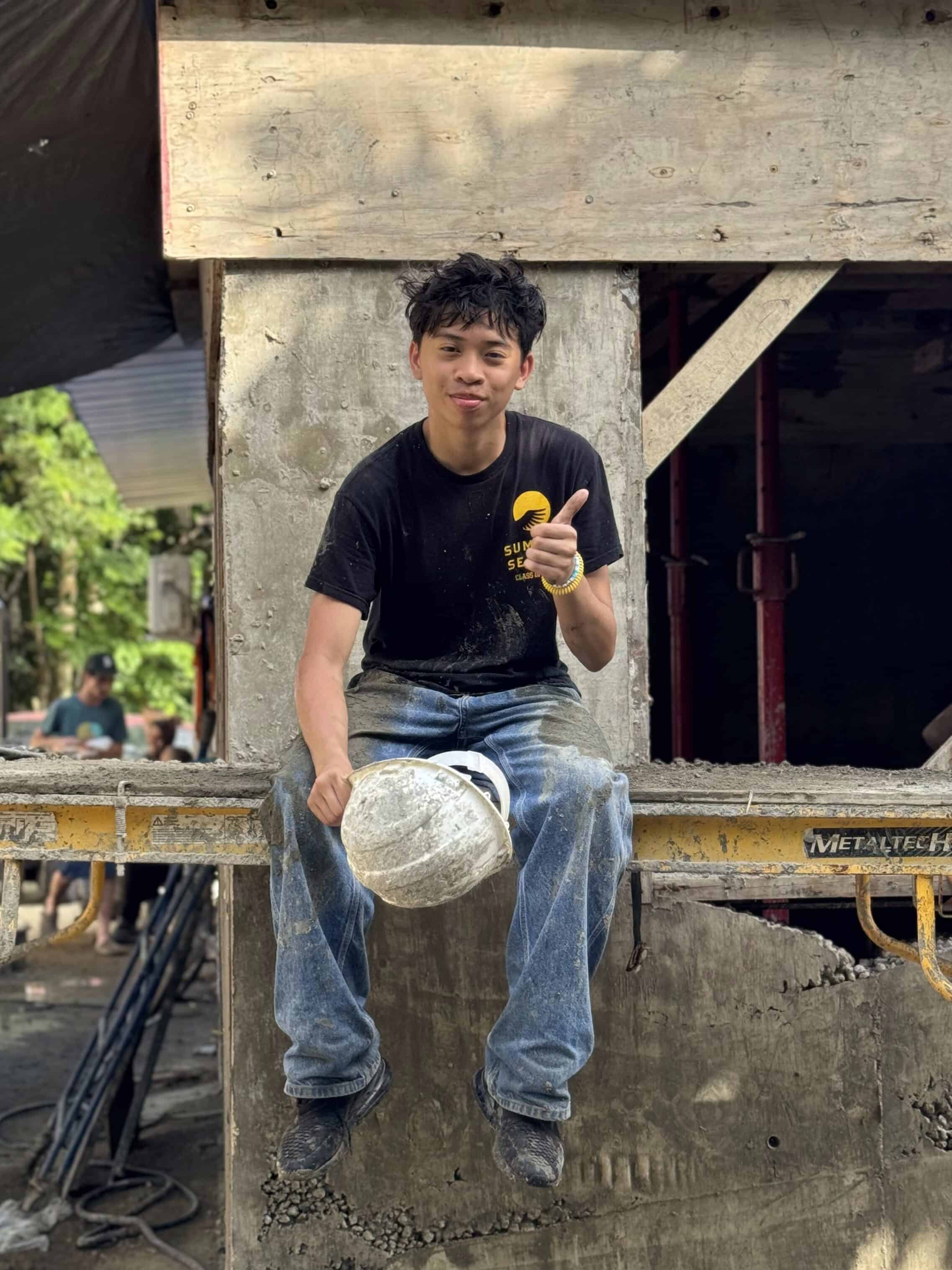 A young man in work clothes sits on a construction beam holding a hard hat and giving a thumbs up, with a building under construction in the background—a perfect scene for tech-free travel programs focused on hands-on experience.
