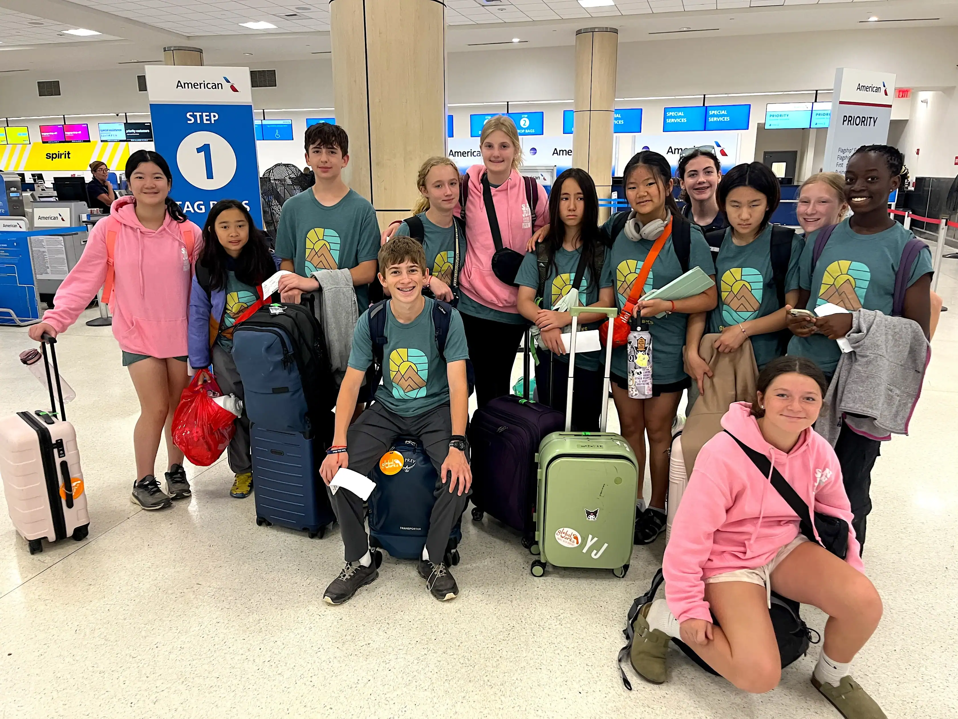 A group of teens with suitcases and backpacks pose together at an airport check-in area, wearing matching t-shirts and pink jackets. Signs for airline check-in are visible in the background.