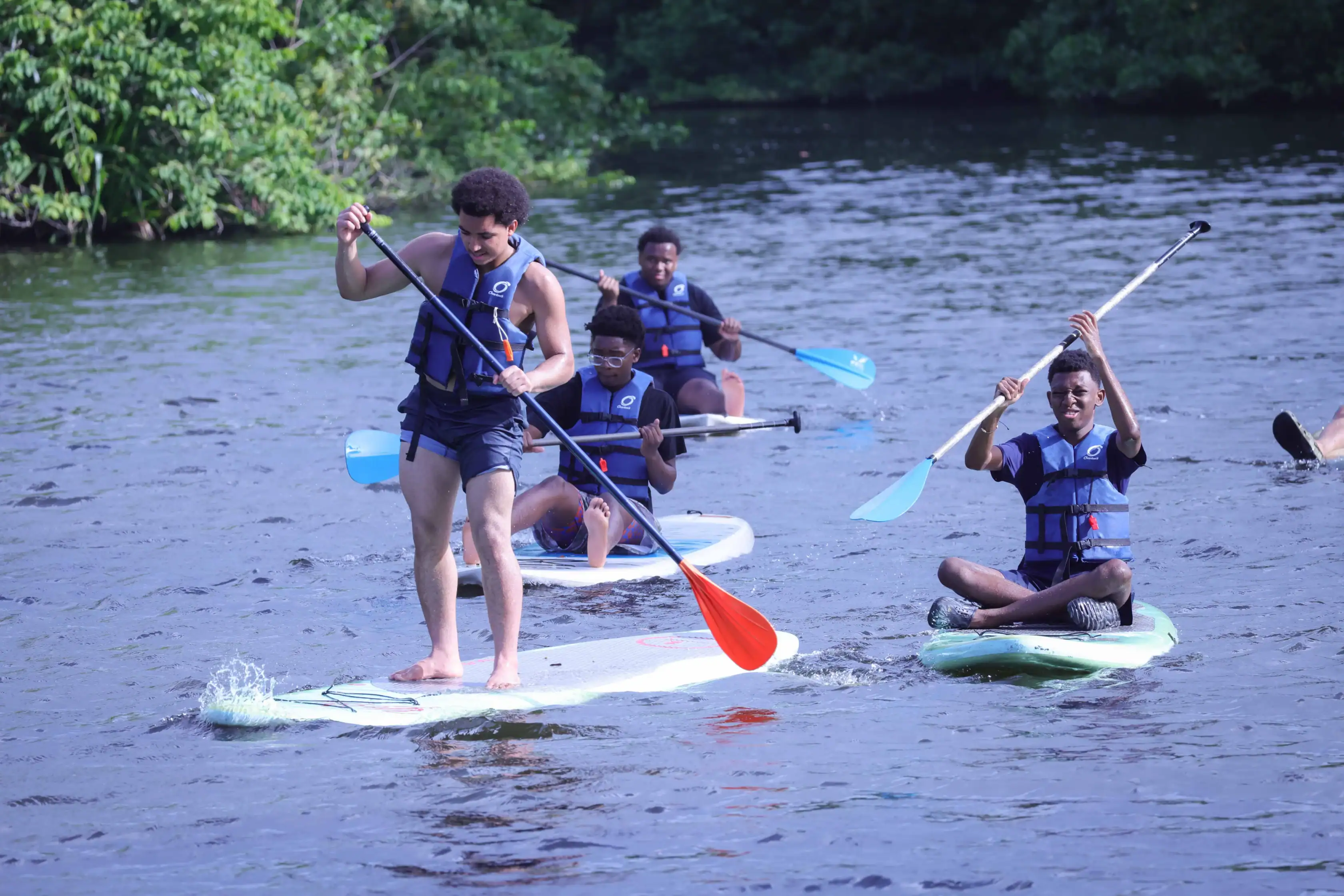 Four people wearing life vests paddle on stand-up paddleboards together on a calm river, surrounded by green trees.