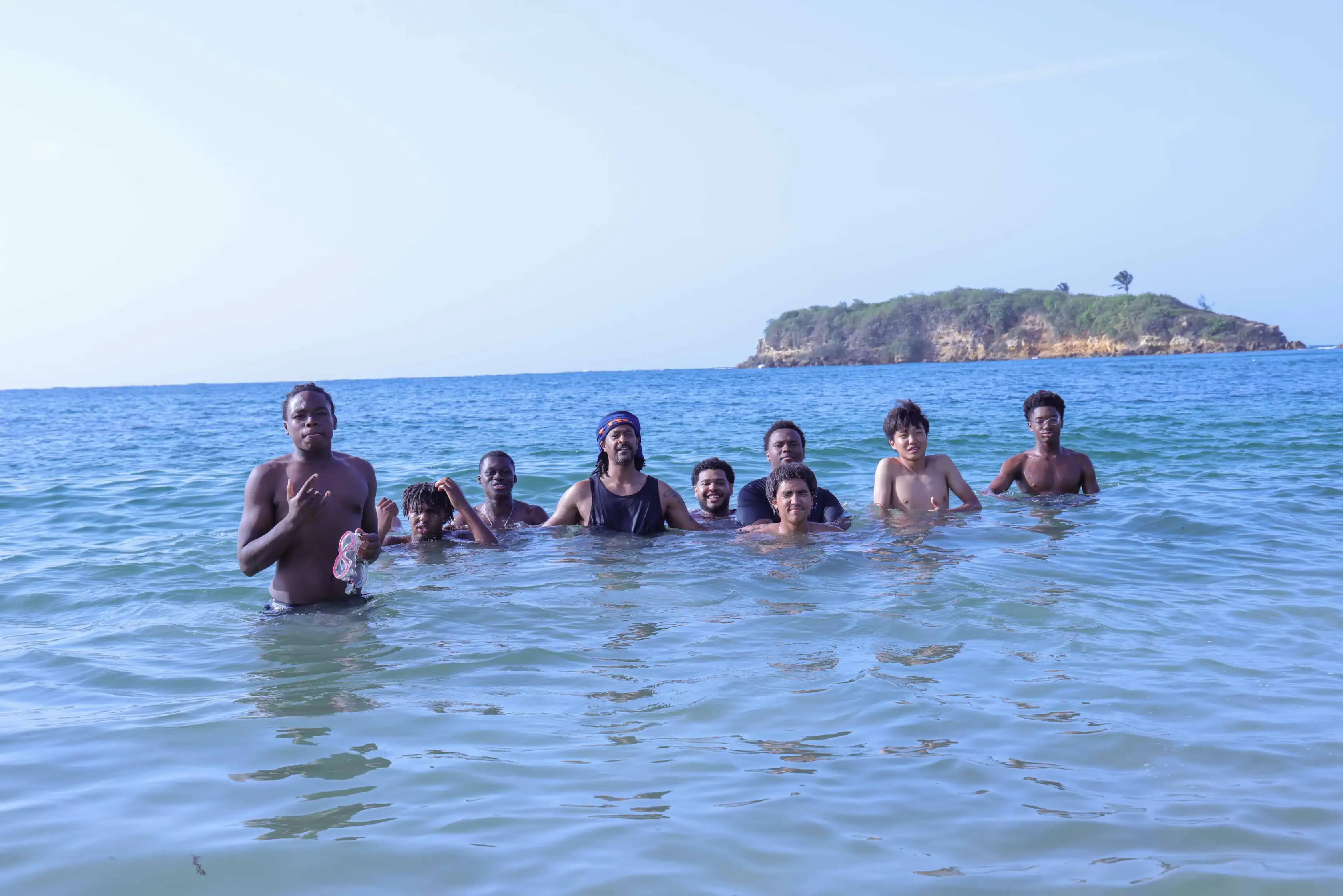 A group of people stand together in the shallow ocean water, with a small island visible in the distance under a clear sky.