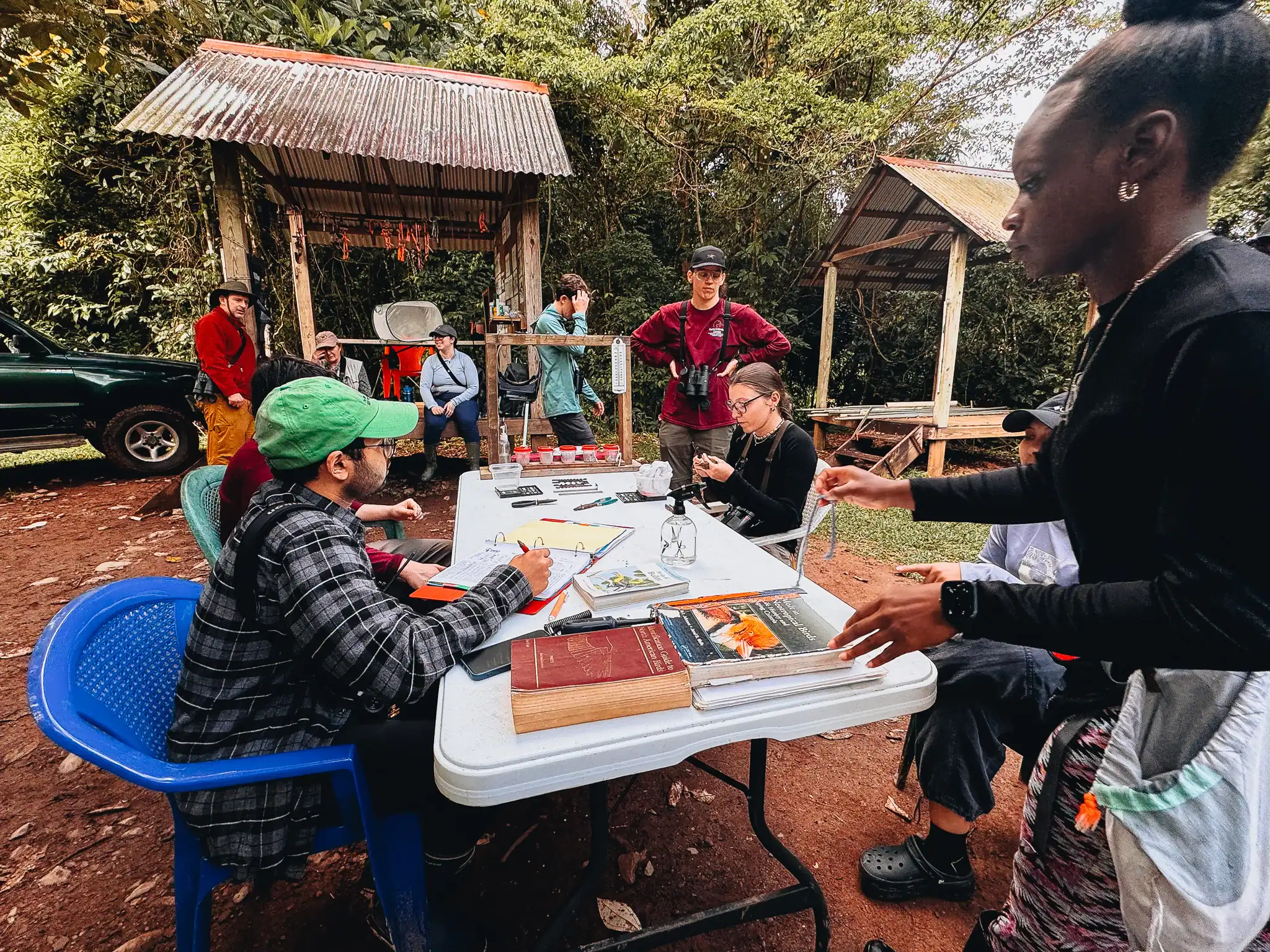 A group of people work at an outdoor table with books and notebooks, surrounded by trees and wooden shelters.