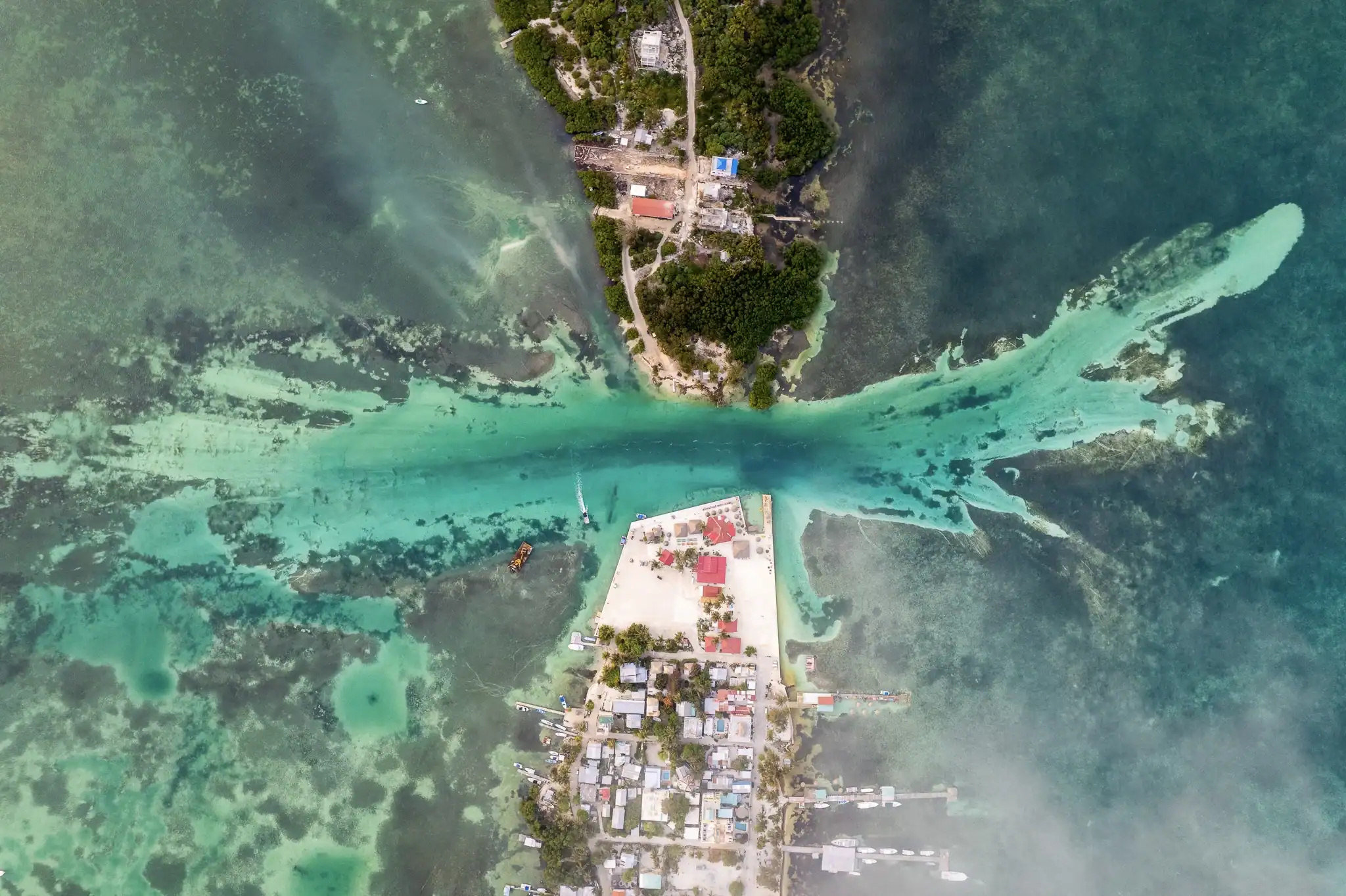 Aerial view of a narrow island split by a turquoise water channel, with buildings and trees on both sides, surrounded by clear, shallow sea.