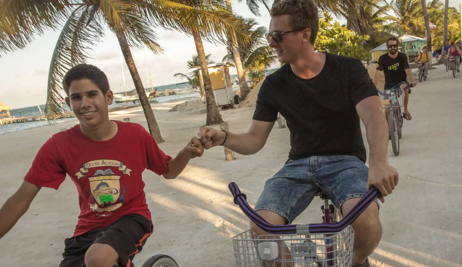 Two young men ride bikes on a sandy path lined with palm trees, holding hands and smiling, with more cyclists and the beach visible in the background.