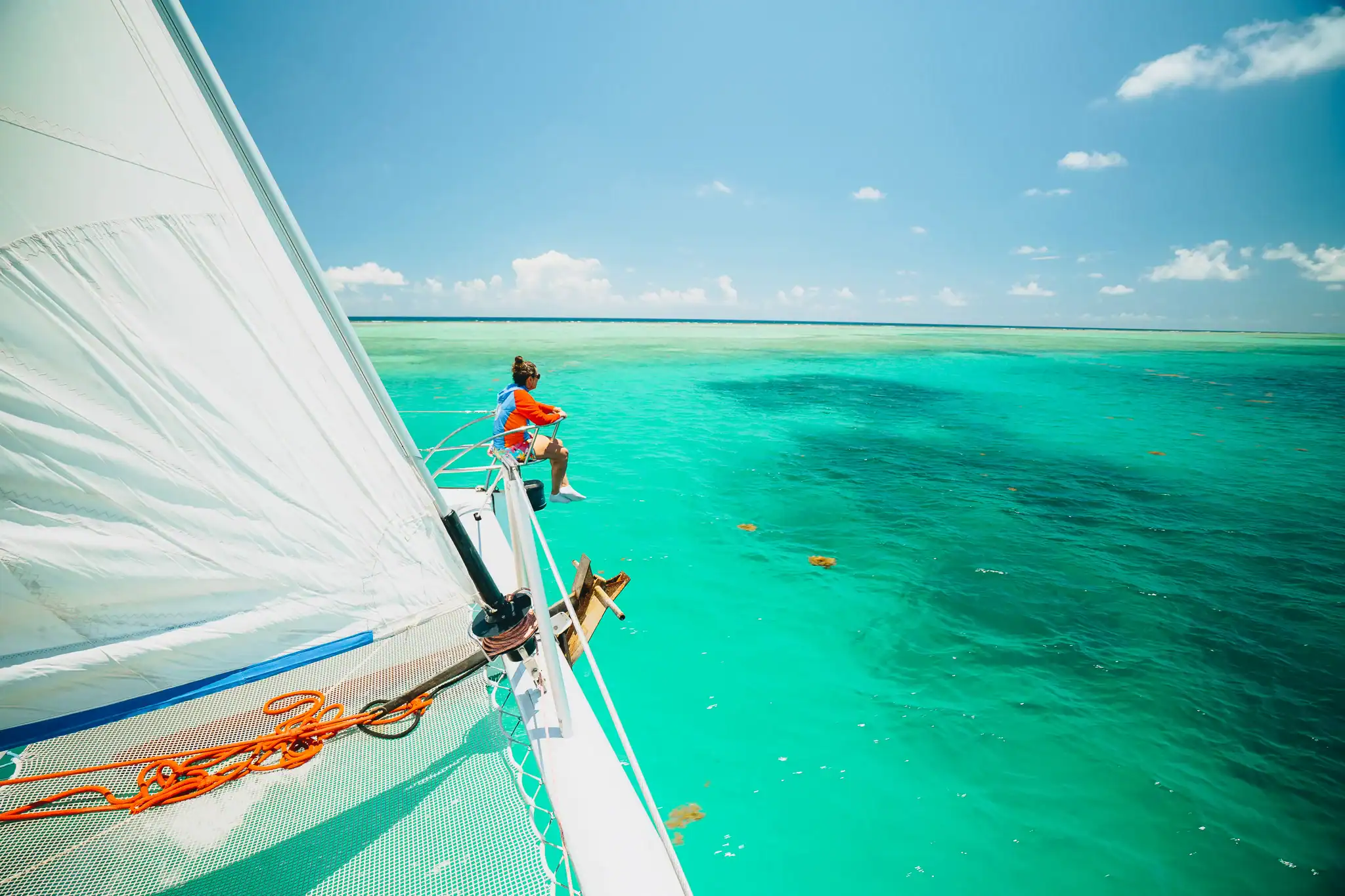 Person sits on the edge of a sailboat floating on clear turquoise water under a sunny sky.