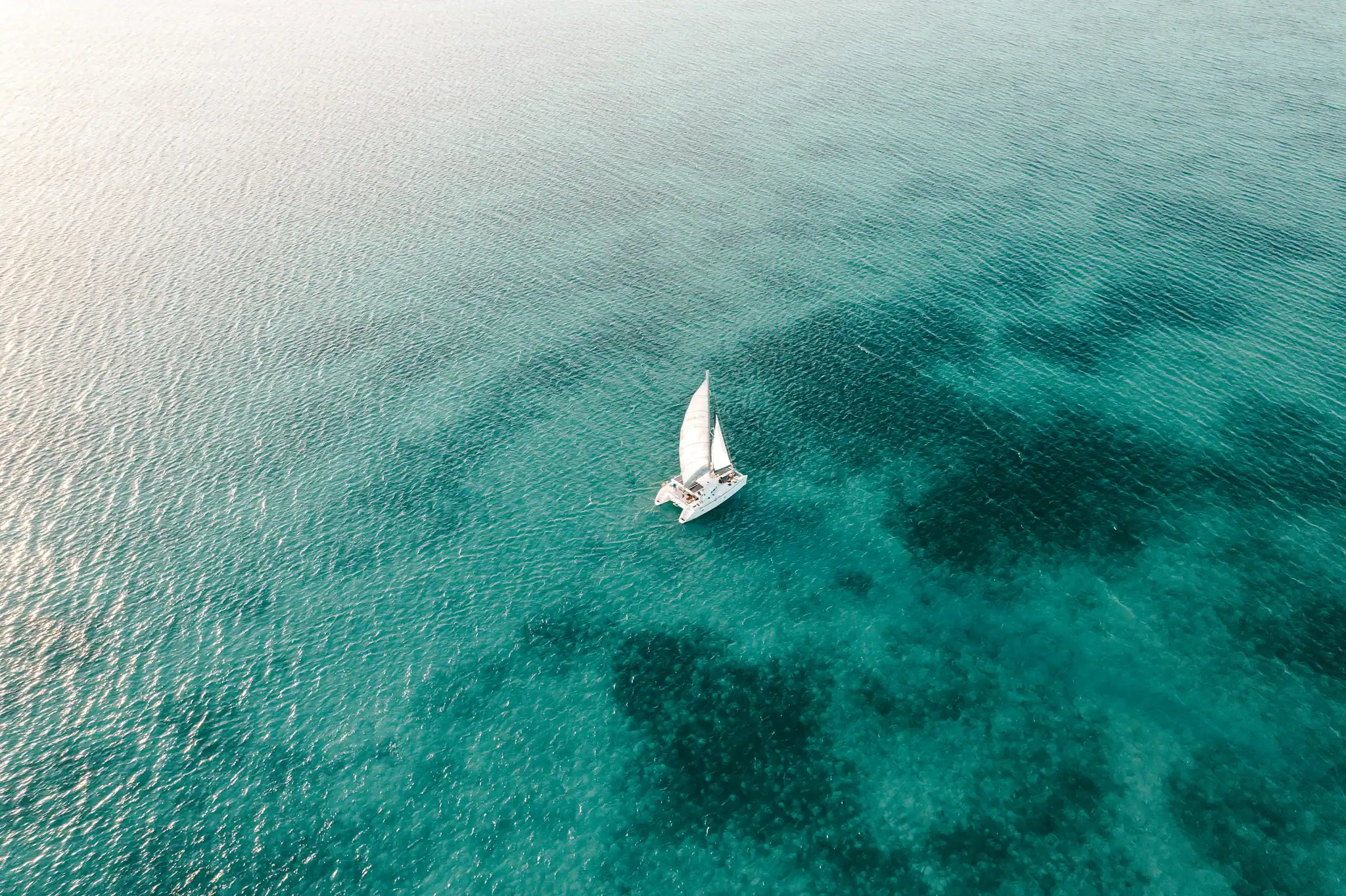 A sailboat with white sails moves across clear turquoise water, viewed from above. The sea appears calm and expansive.