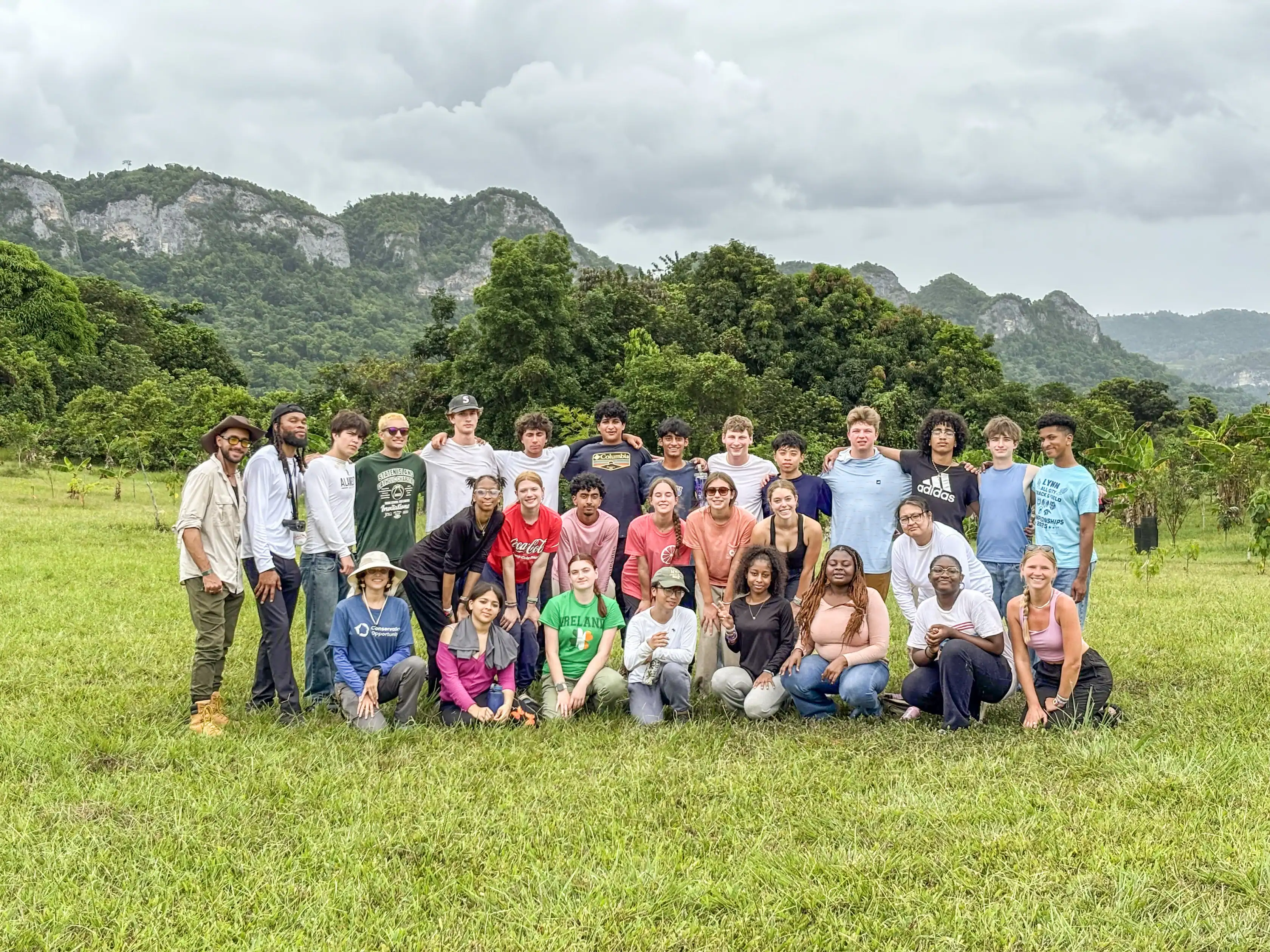 A group of people poses together outdoors on grass with green trees and rocky hills in the background under a cloudy sky.
