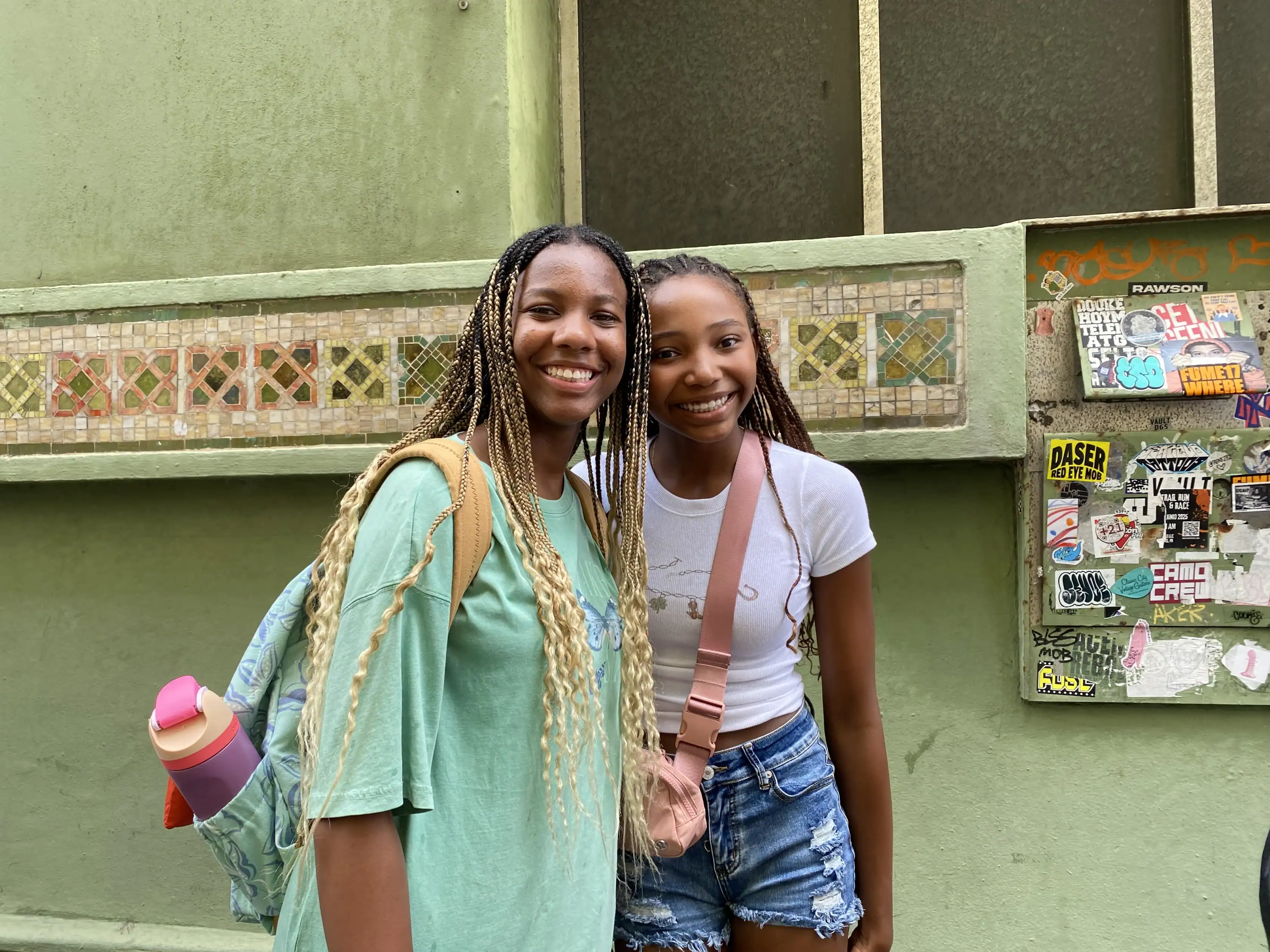 Two young people stand smiling in front of a green wall covered with tiles and stickers. One wears a green shirt with a backpack; the other wears a white top and denim shorts.