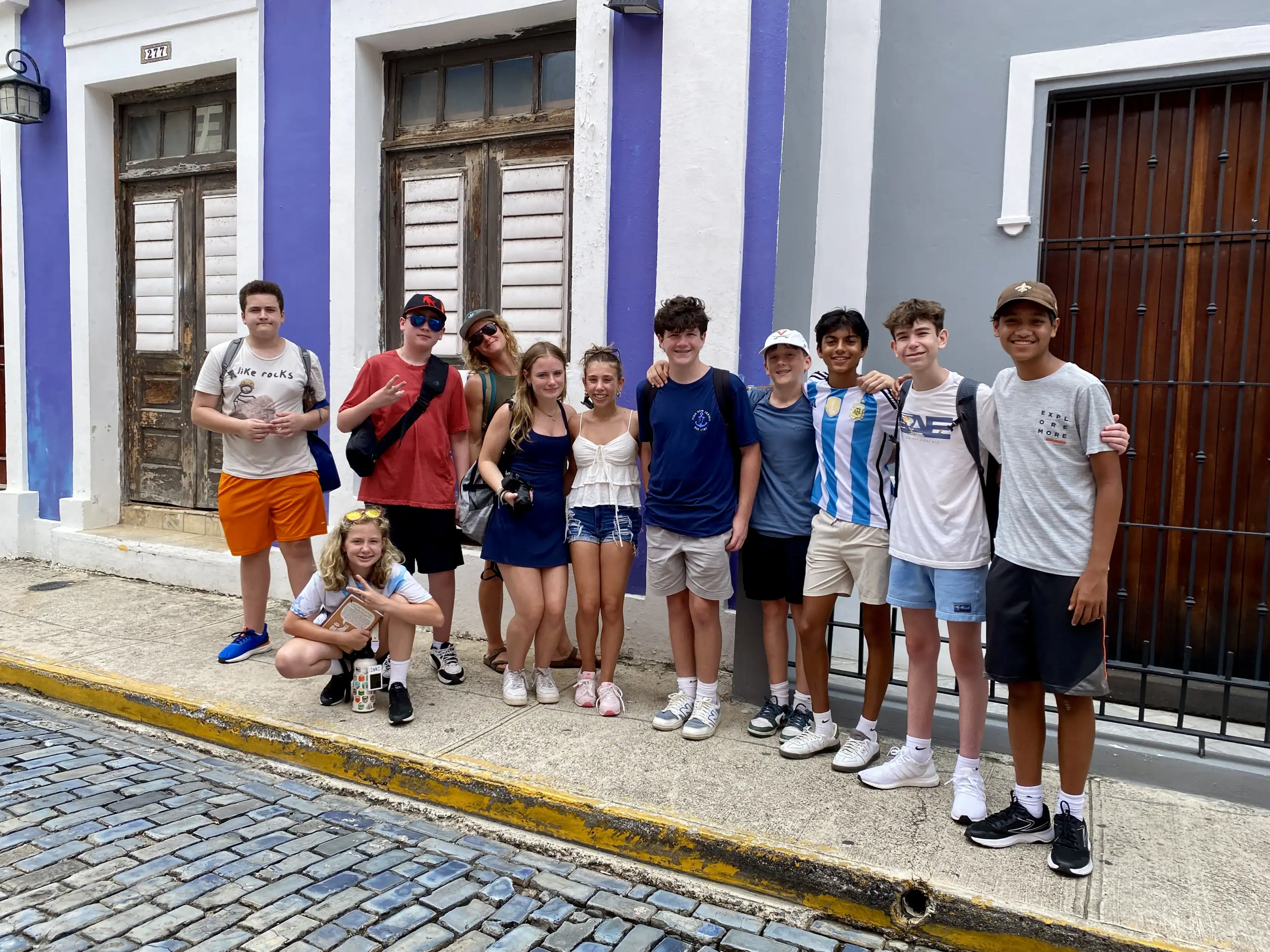 A group of teenagers poses together on a sidewalk in front of colorful buildings with wooden doors and windows.