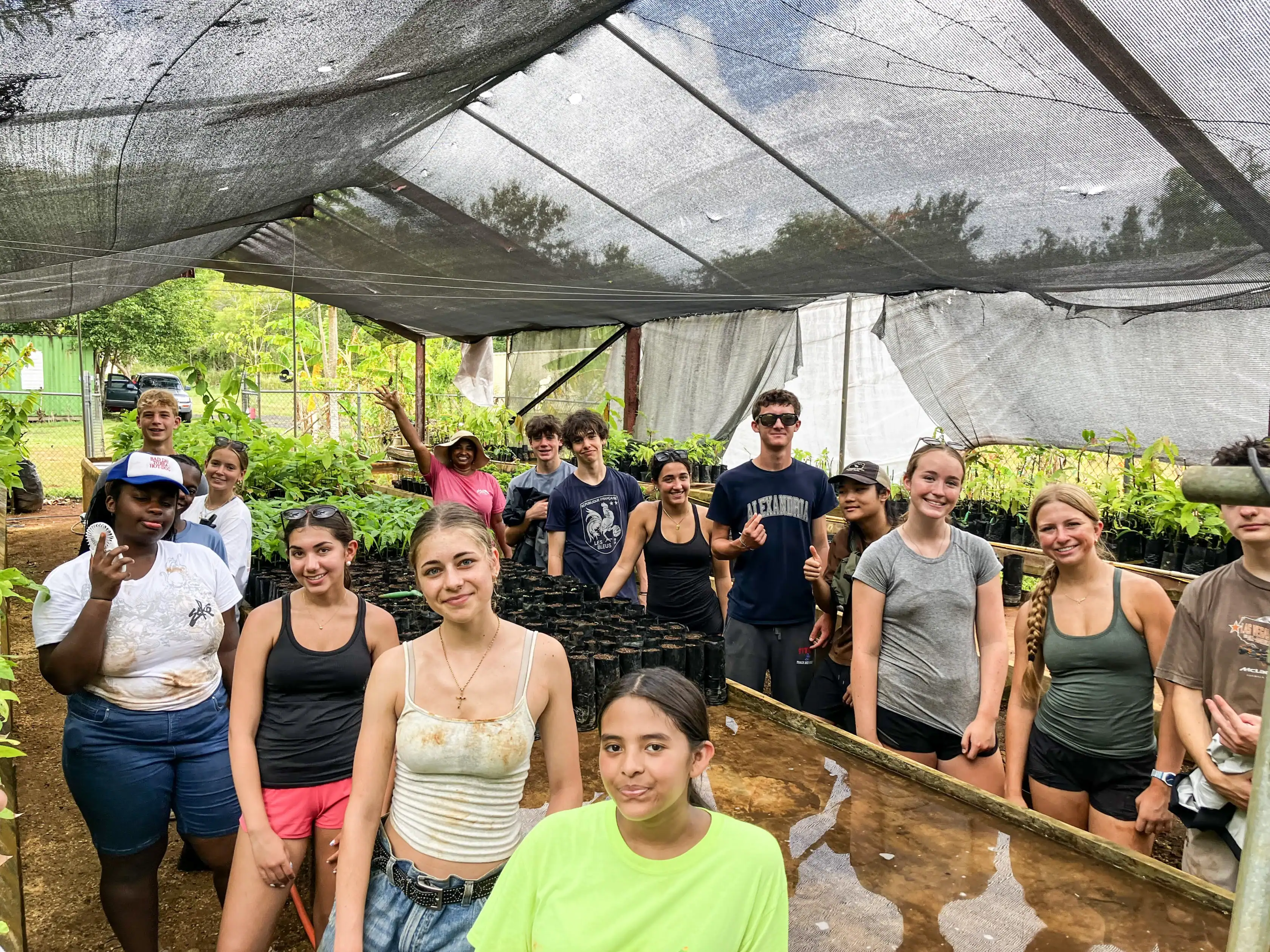A group of people pose in a greenhouse, surrounded by plants and pots, wearing casual outdoor clothing, some smiling and some making gestures, with shade cloth overhead.