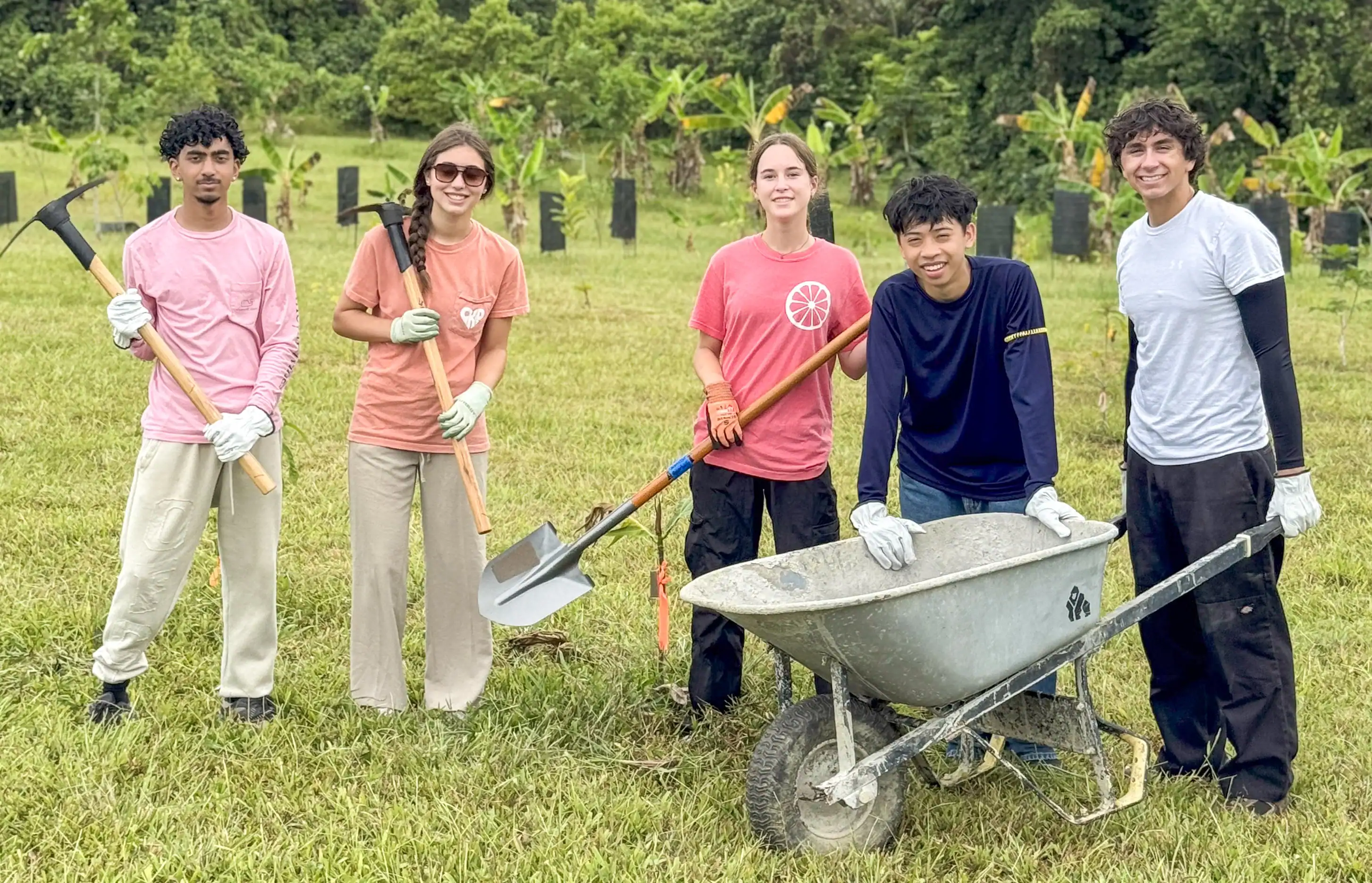 Five people stand outdoors on a grassy field with a wheelbarrow and gardening tools, dressed casually for a planting or gardening activity. Trees and plants are visible in the background.