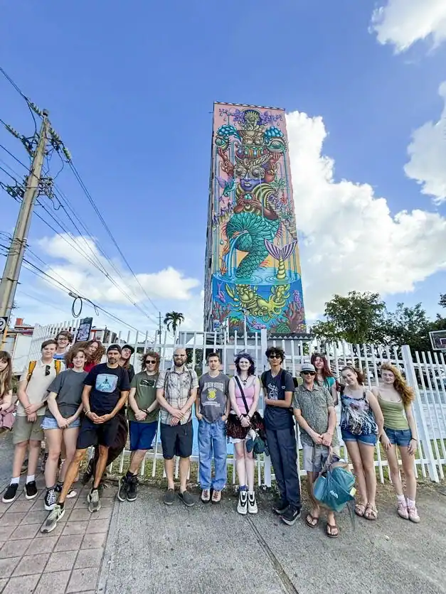 A group of people stands in front of a tall, colorful mural on a building, with a blue sky and clouds in the background.