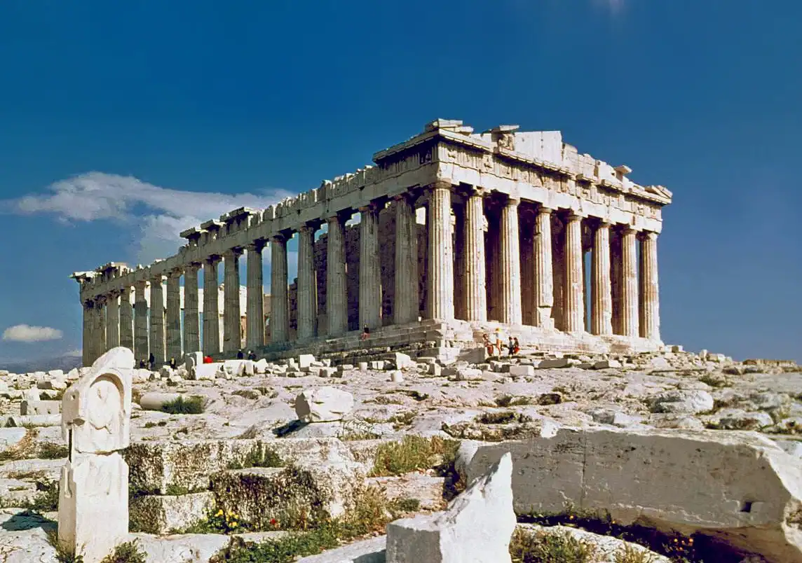 The Parthenon, an ancient Greek temple with Doric columns, stands on the Acropolis of Athens under a clear blue sky, surrounded by scattered stone ruins.