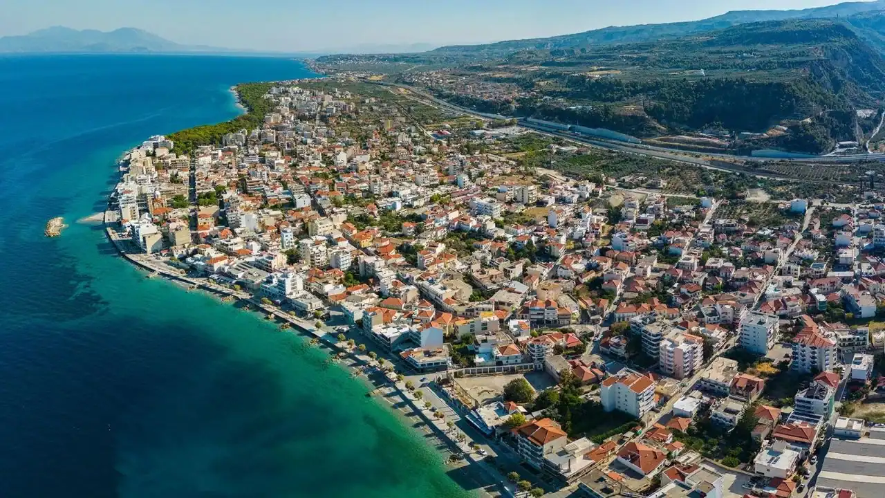 Aerial view of a coastal town with densely packed buildings, clear turquoise water, and a road running parallel to the shoreline, with mountains in the background.