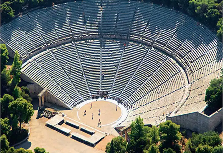 Aerial view of a large, ancient, stone amphitheater surrounded by trees with a circular stage and tiered seating.