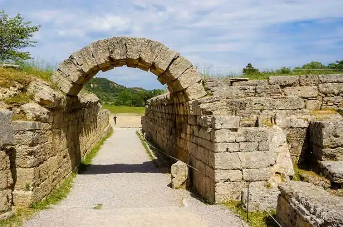 Stone archway at an ancient ruin site with gravel pathway leading through it, surrounded by stone walls and greenery under a partly cloudy sky.