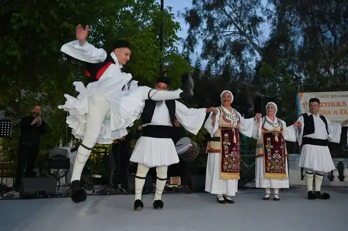 People in traditional Greek costumes perform a folk dance on an outdoor stage; one dancer is mid-air in a high kick, while others hold hands in a line.