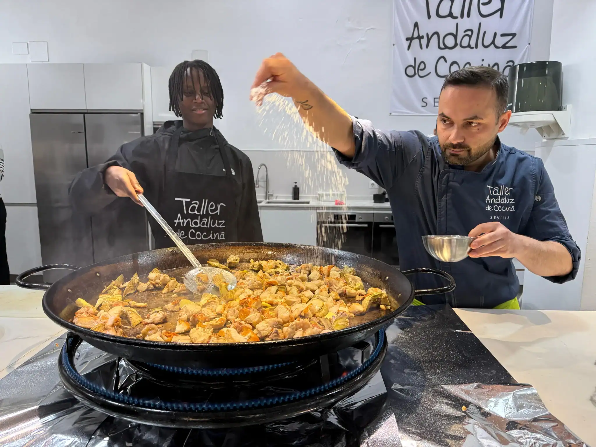 Two chefs in branded aprons cooking chicken and artichokes in a large paella pan over a gas burner.
