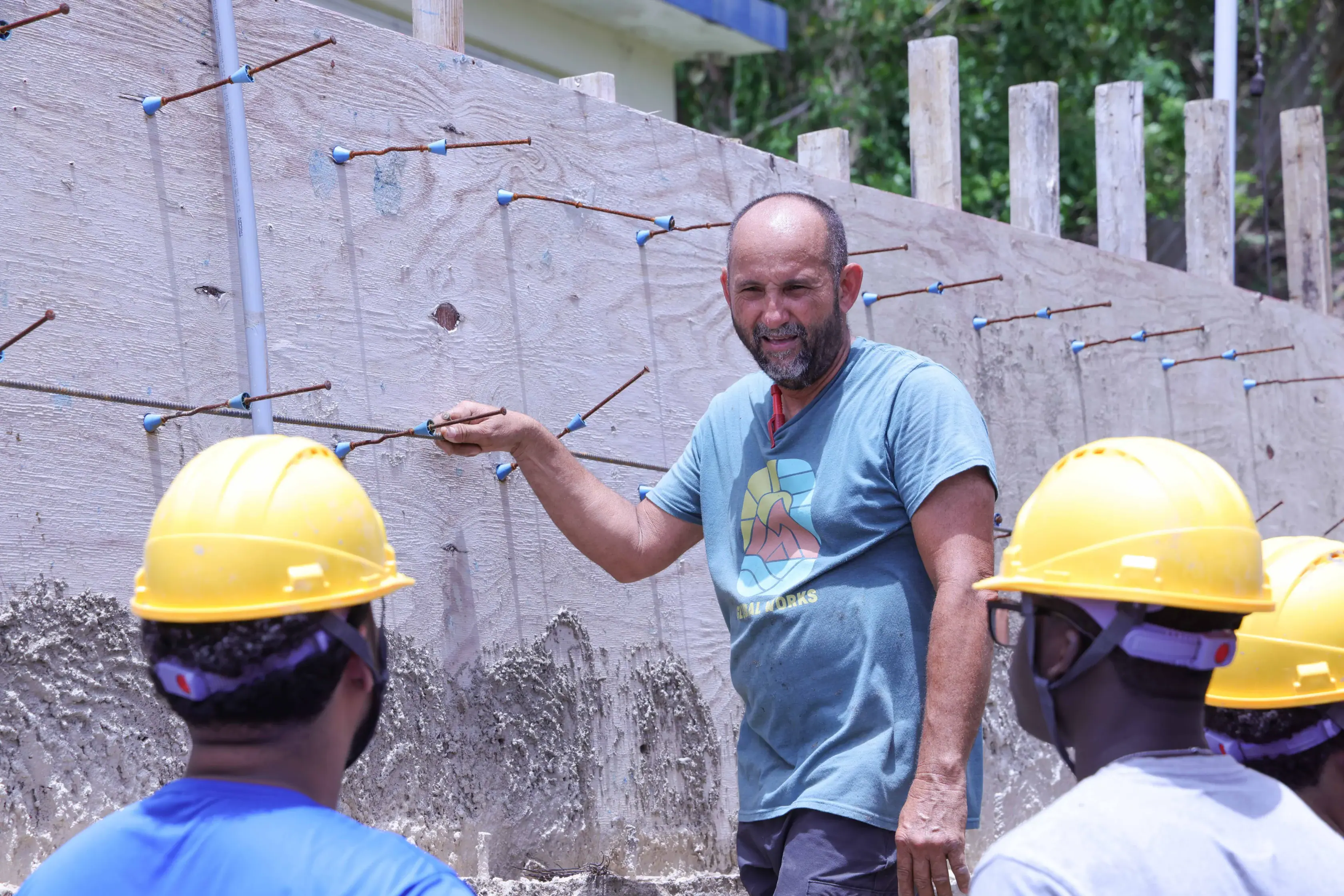 A man in a graphic t-shirt gestures toward a plywood concrete form braced with metal rebar, while two construction workers in yellow hard hats look on.