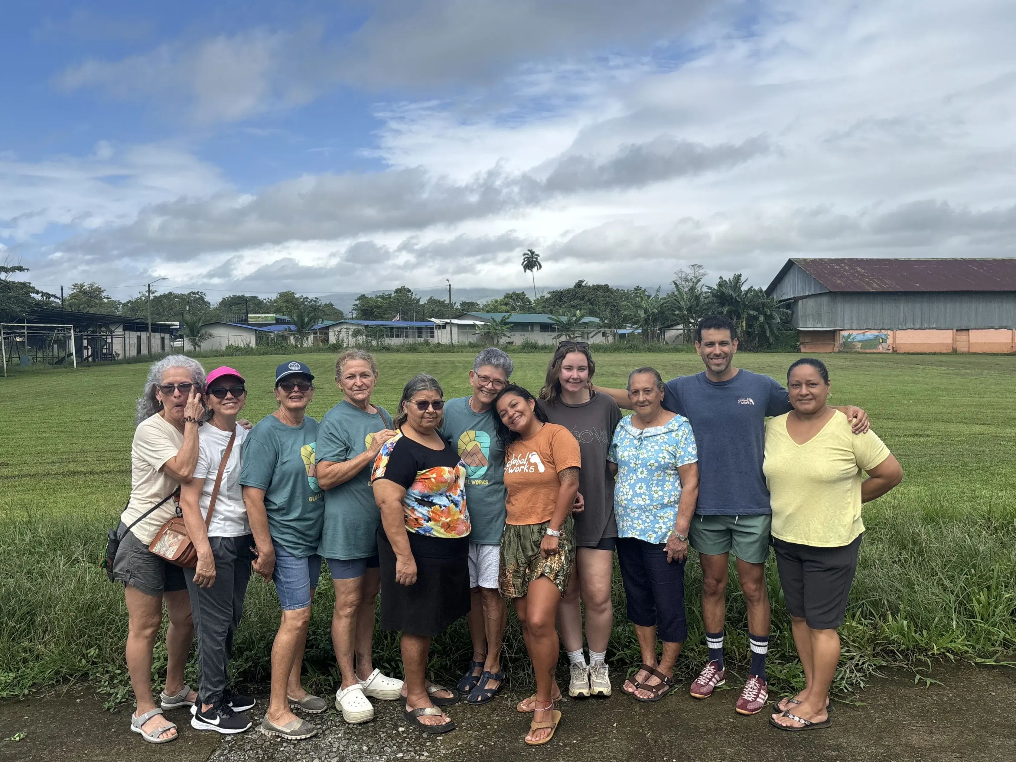 A group of eleven diverse people stand in a line on a paved path in front of a grassy field.