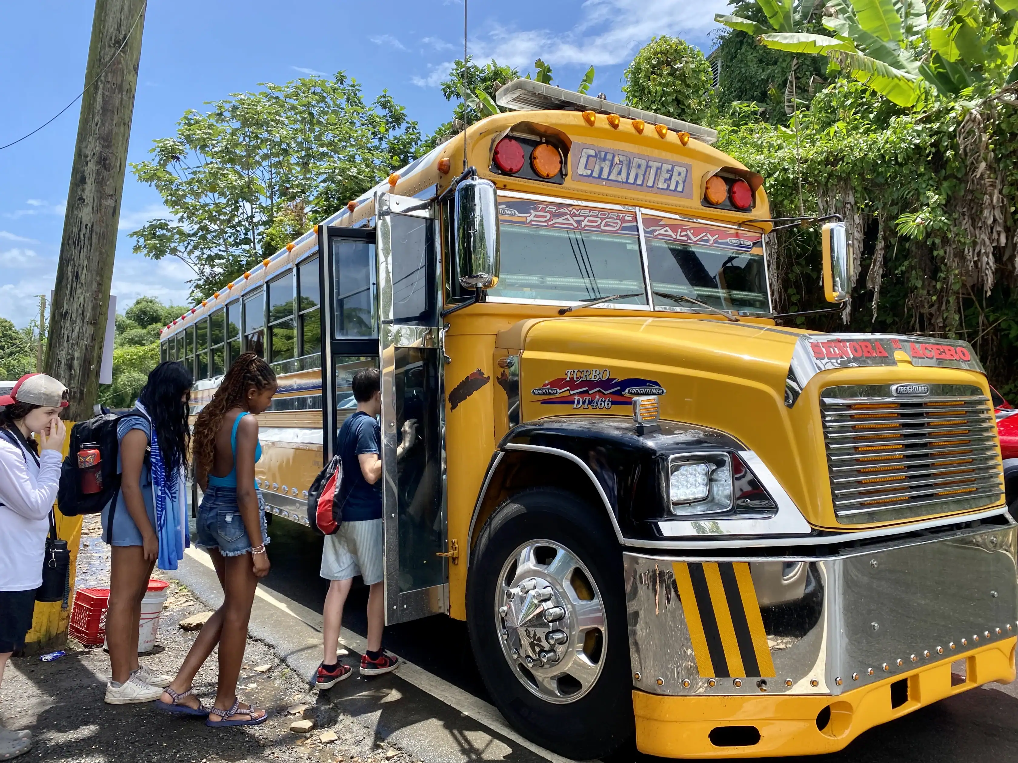 A group of people board a yellow Freightliner charter bus parked on the side of a road.