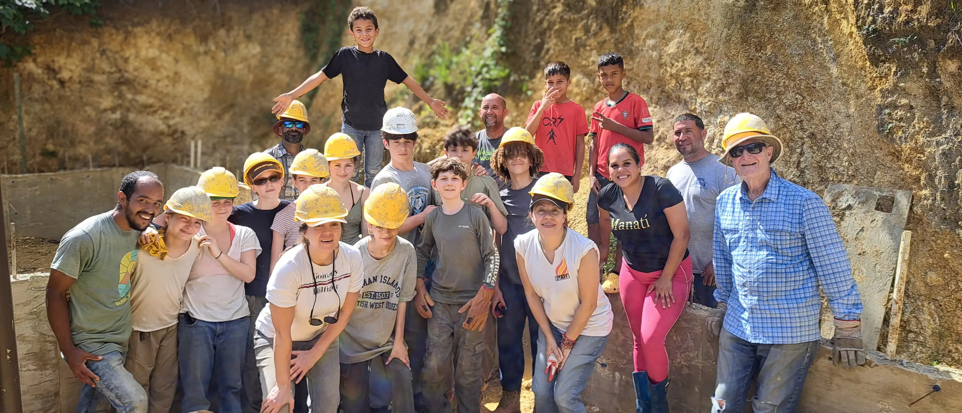A diverse group of people, many wearing yellow hard hats, standing together at a construction site in front of an earthen wall.