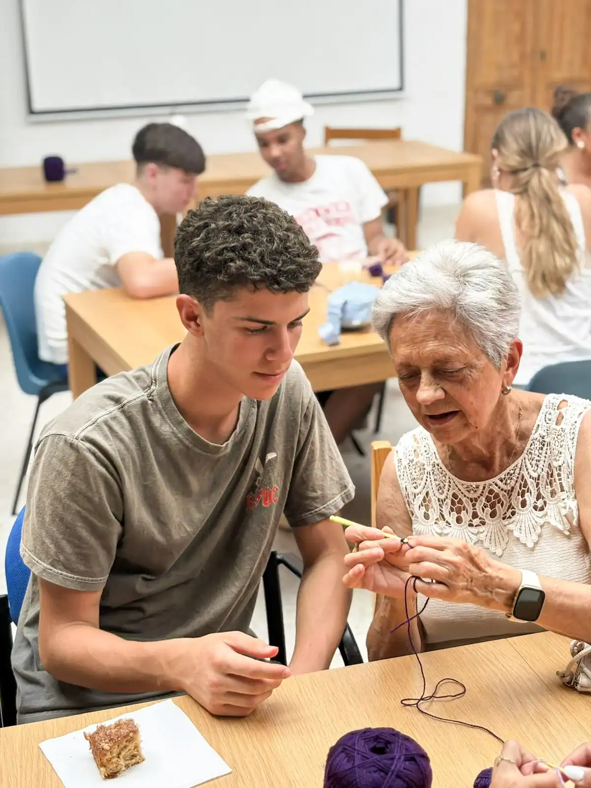 An older woman with short gray hair teaches a young man how to crochet with dark purple yarn.