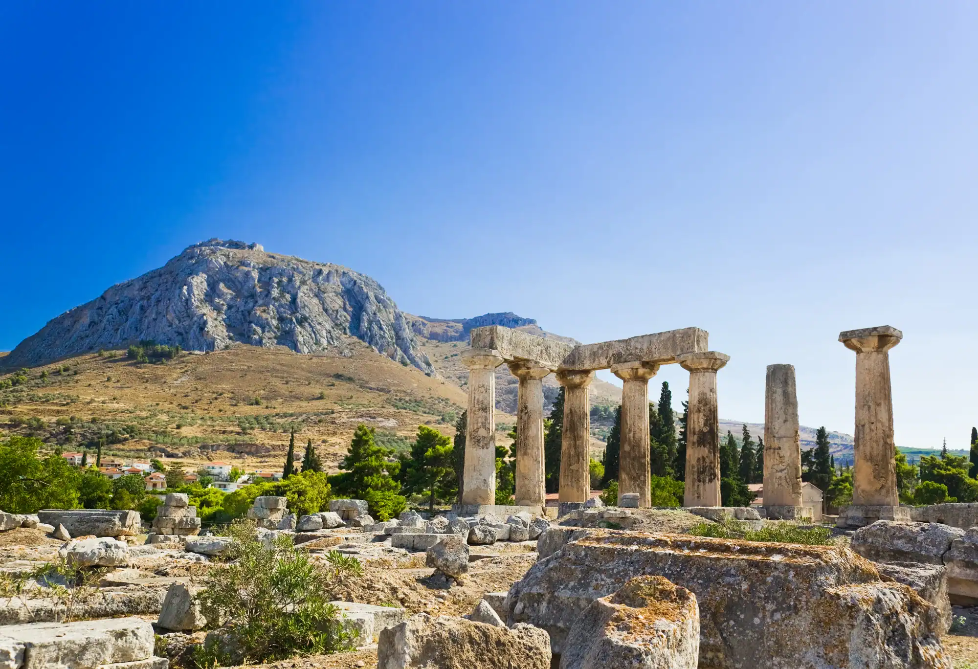 Ancient stone columns of a ruined temple stand in front of a mountain under a clear blue sky, with scattered stones and sparse greenery in the foreground.